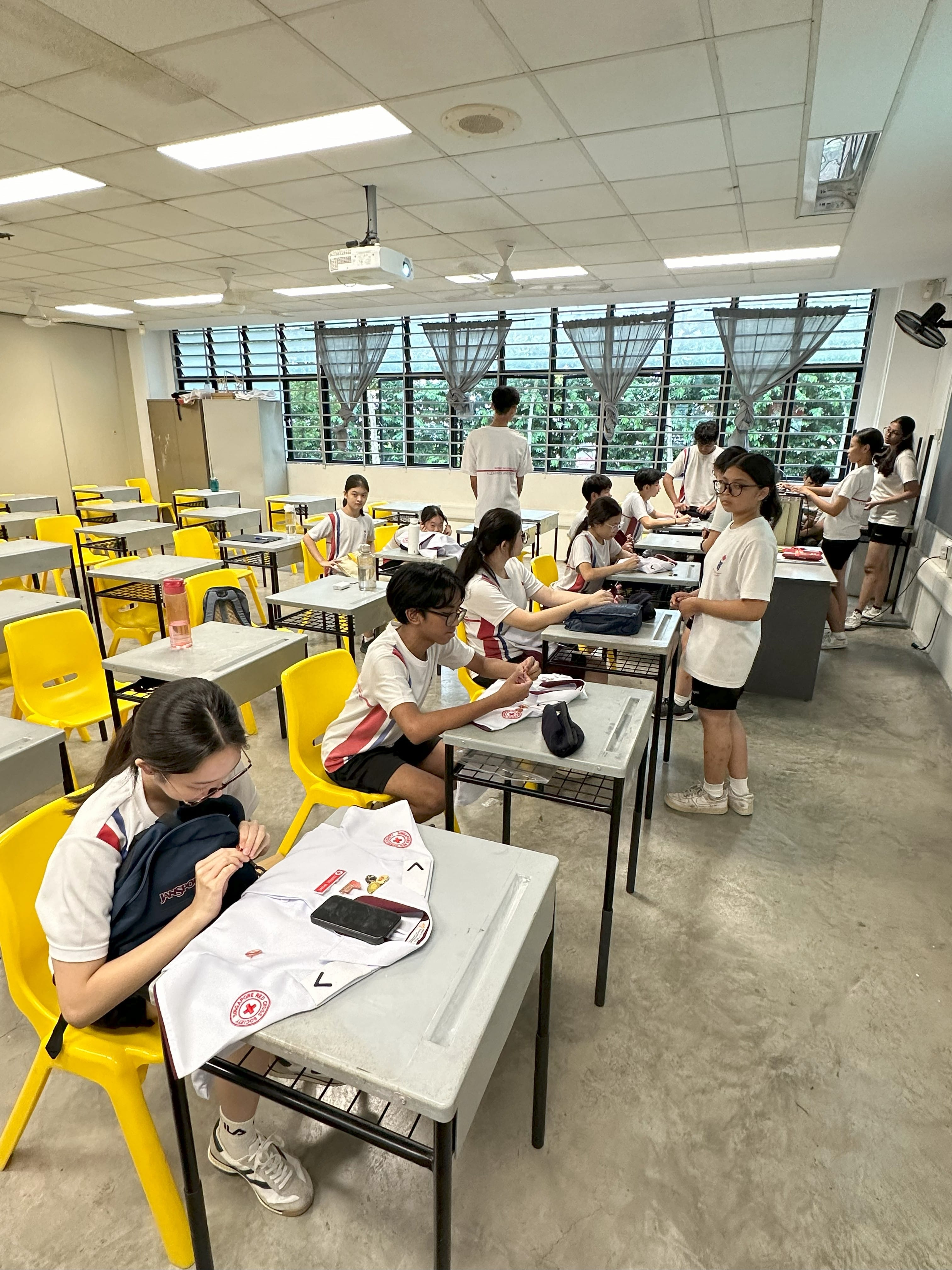 Students in white uniforms sit and stand around desks with yellow chairs in a bright classroom.