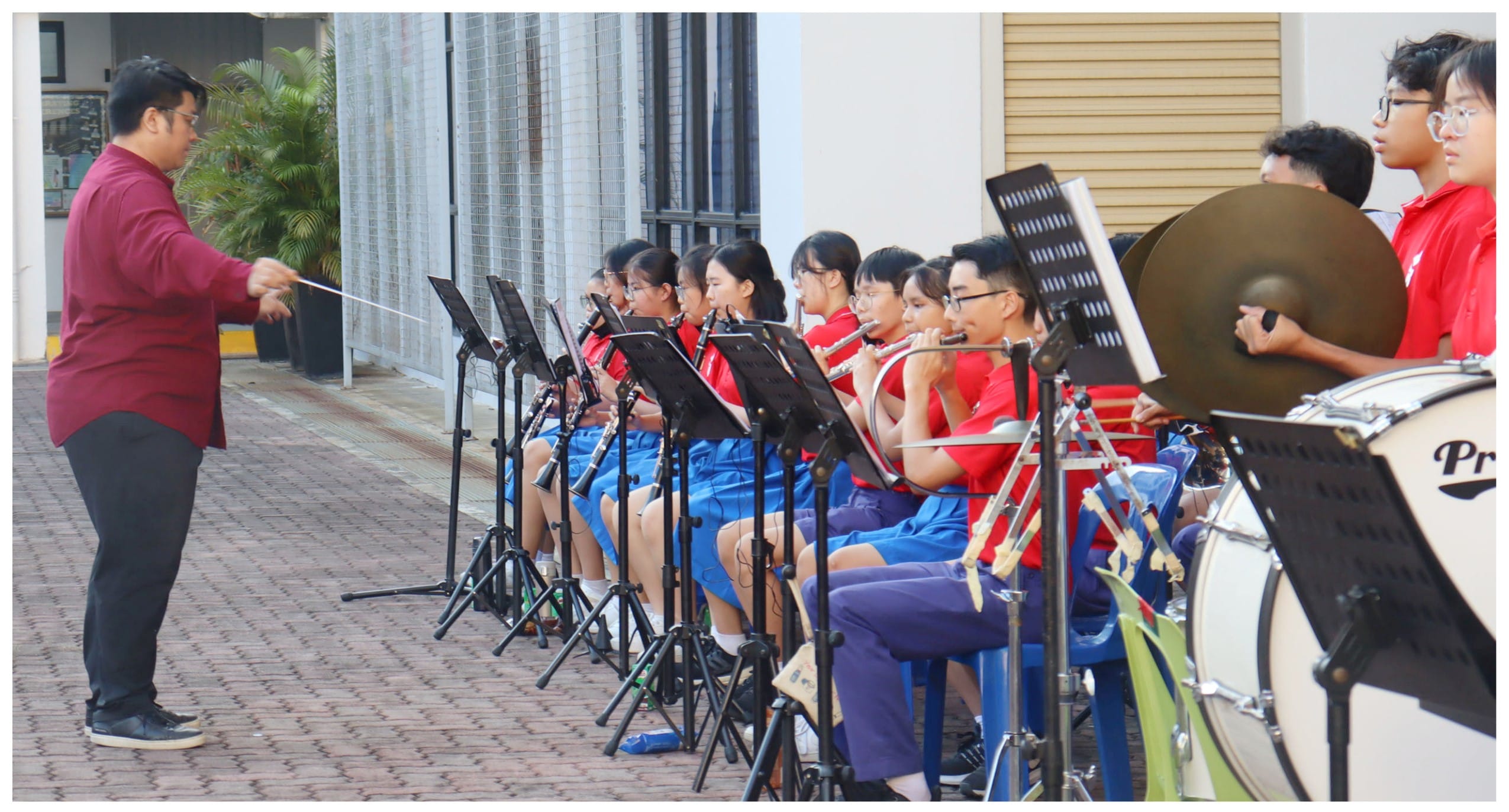 A conductor leads a youth band playing wind instruments and drums outdoors, students in red and blue uniforms with music stands.