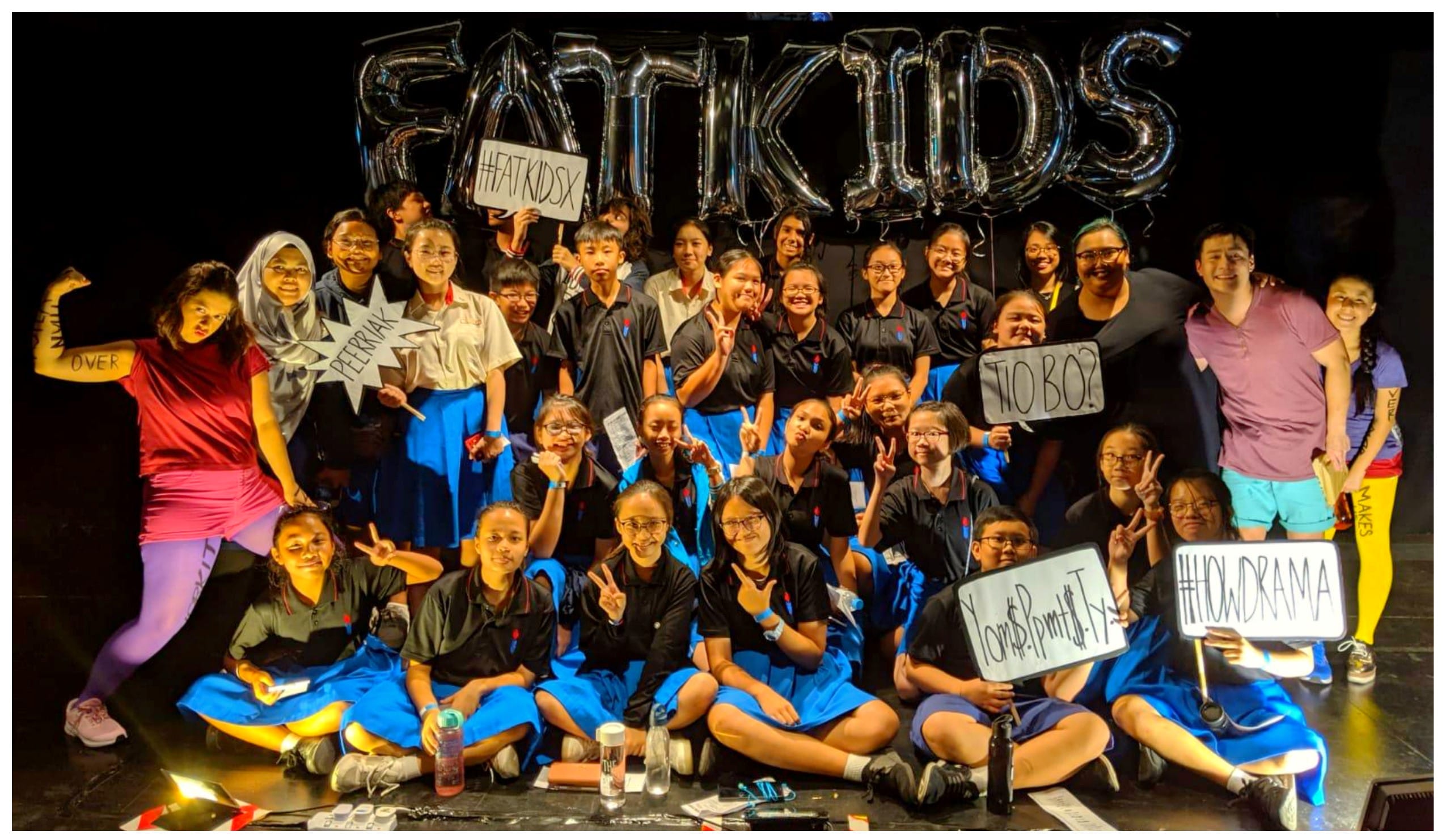 A large group of young students pose on a dark stage under silver "FATKIDS" balloons, holding signs with hashtags.