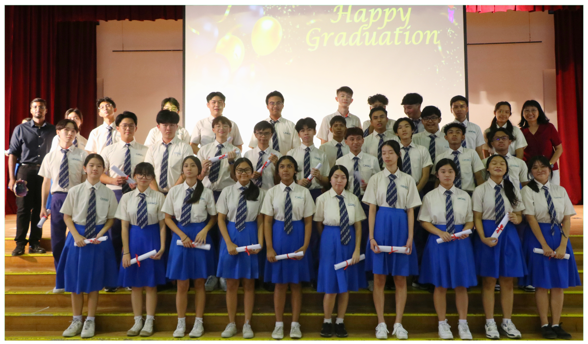 Students in white uniforms with blue ties pose on stage holding diplomas in front of a Happy Graduation screen.