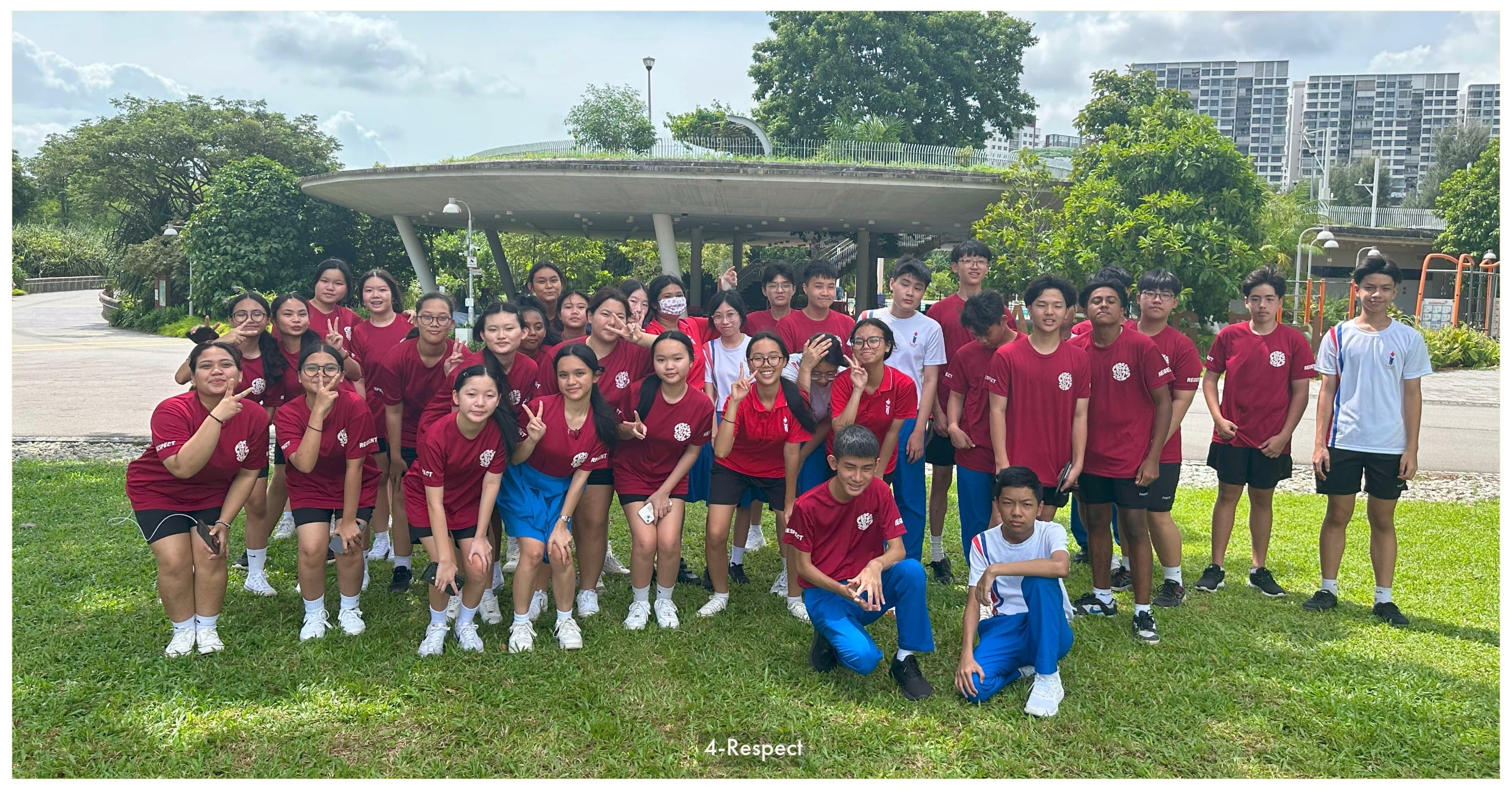 A large group of students in red uniforms posing outdoors on grass near a round pavilion structure, many making peace signs.