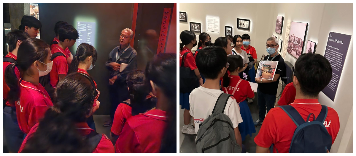 Students in red uniforms listen to a guide during a museum tour, with framed photos and exhibit panels on the walls.