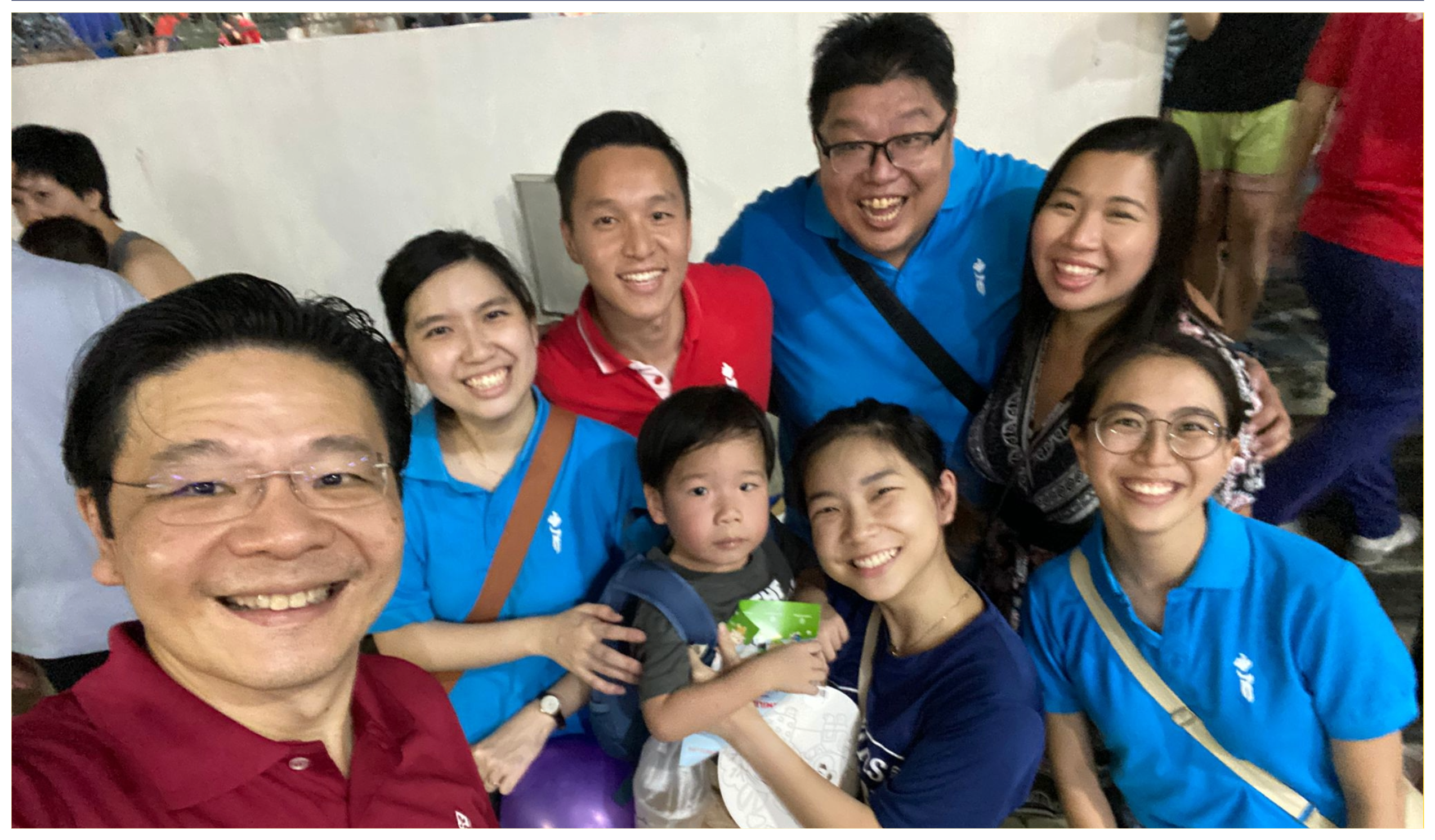 Group selfie of seven smiling adults and a child indoors, with several wearing blue shirts.