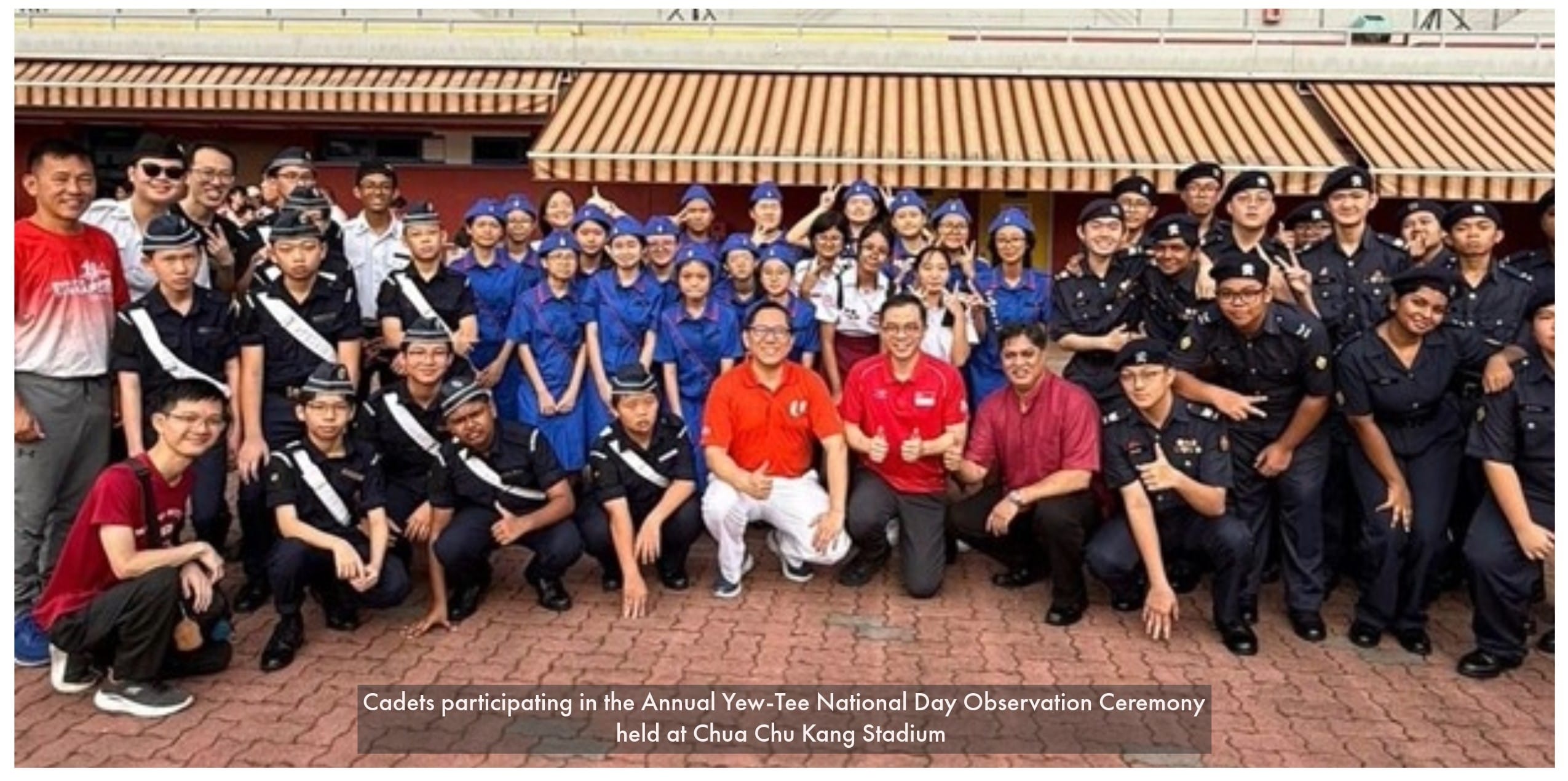 A large group of cadets in uniform pose for a photo with three adults in red shirts outdoors.