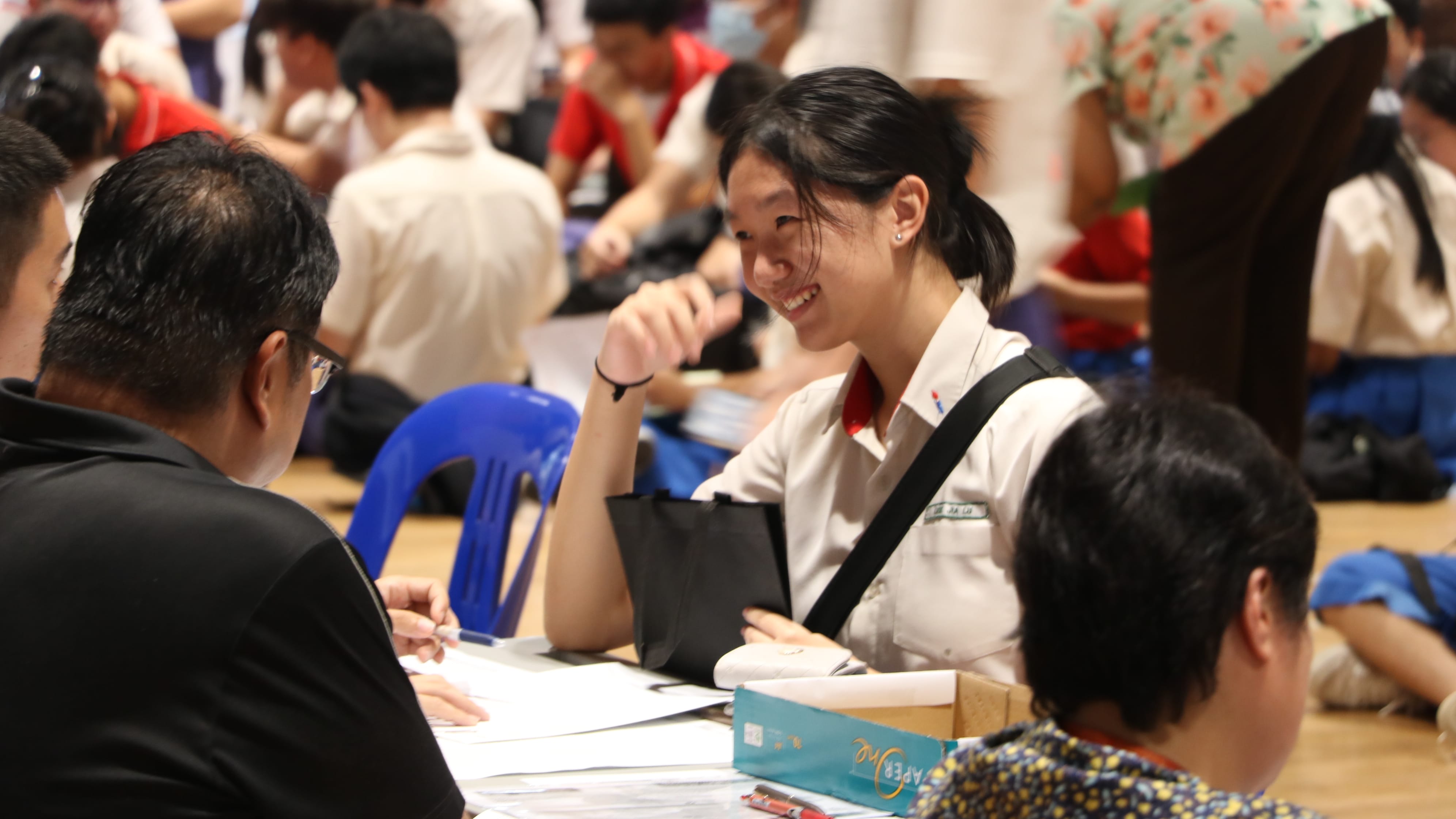 A smiling young woman in a white uniform sits at a table with papers, talking to a man in a crowded hall.