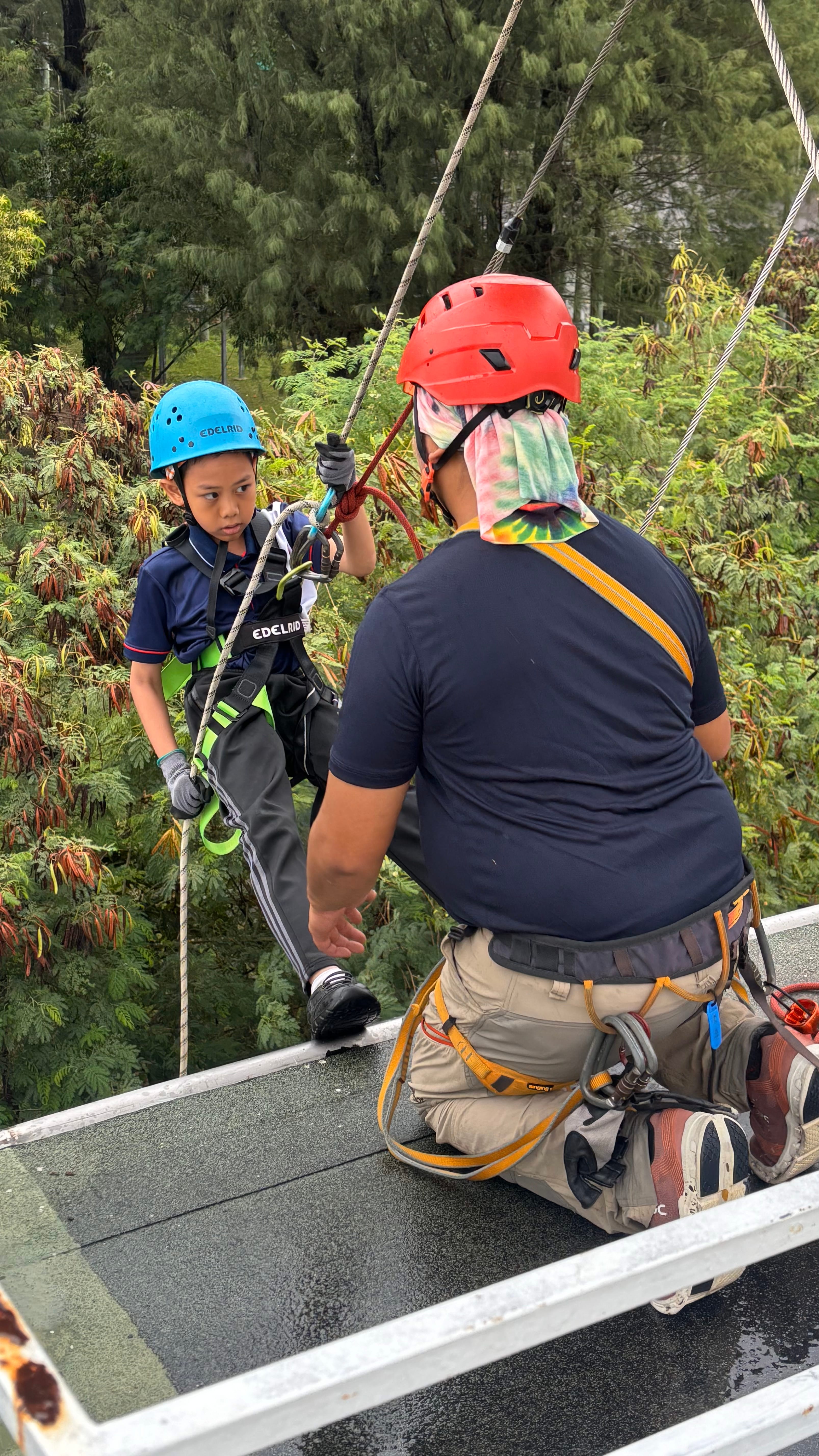 Child in blue helmet clipped to ropes while an instructor in red helmet kneels on a platform amid trees.