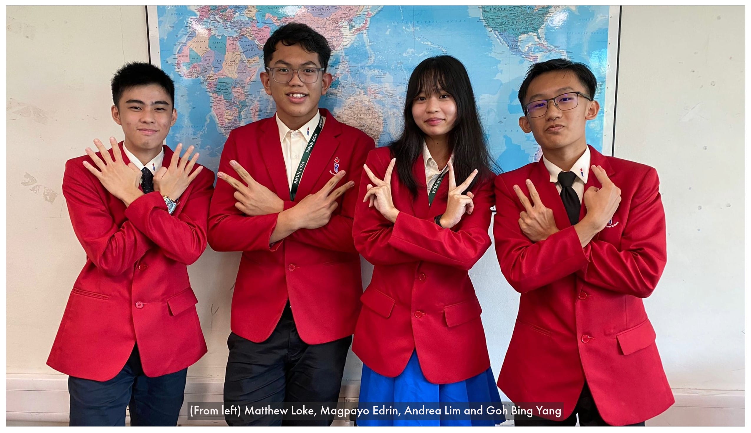 Four students in red blazers pose with hand gestures in front of a world map on a wall.