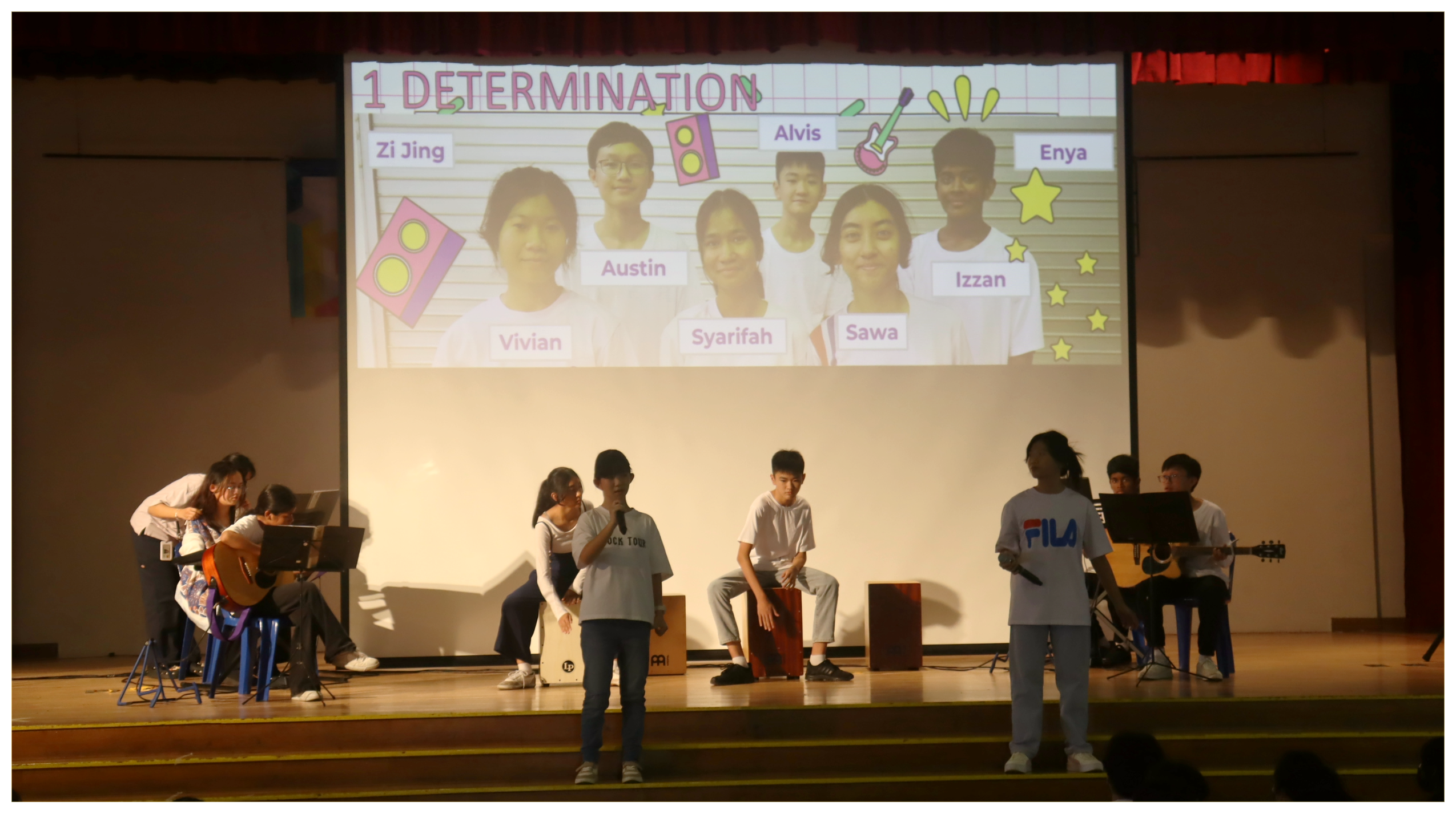 Students perform on stage with guitars and drums beneath a projection titled 1 Determination with names.