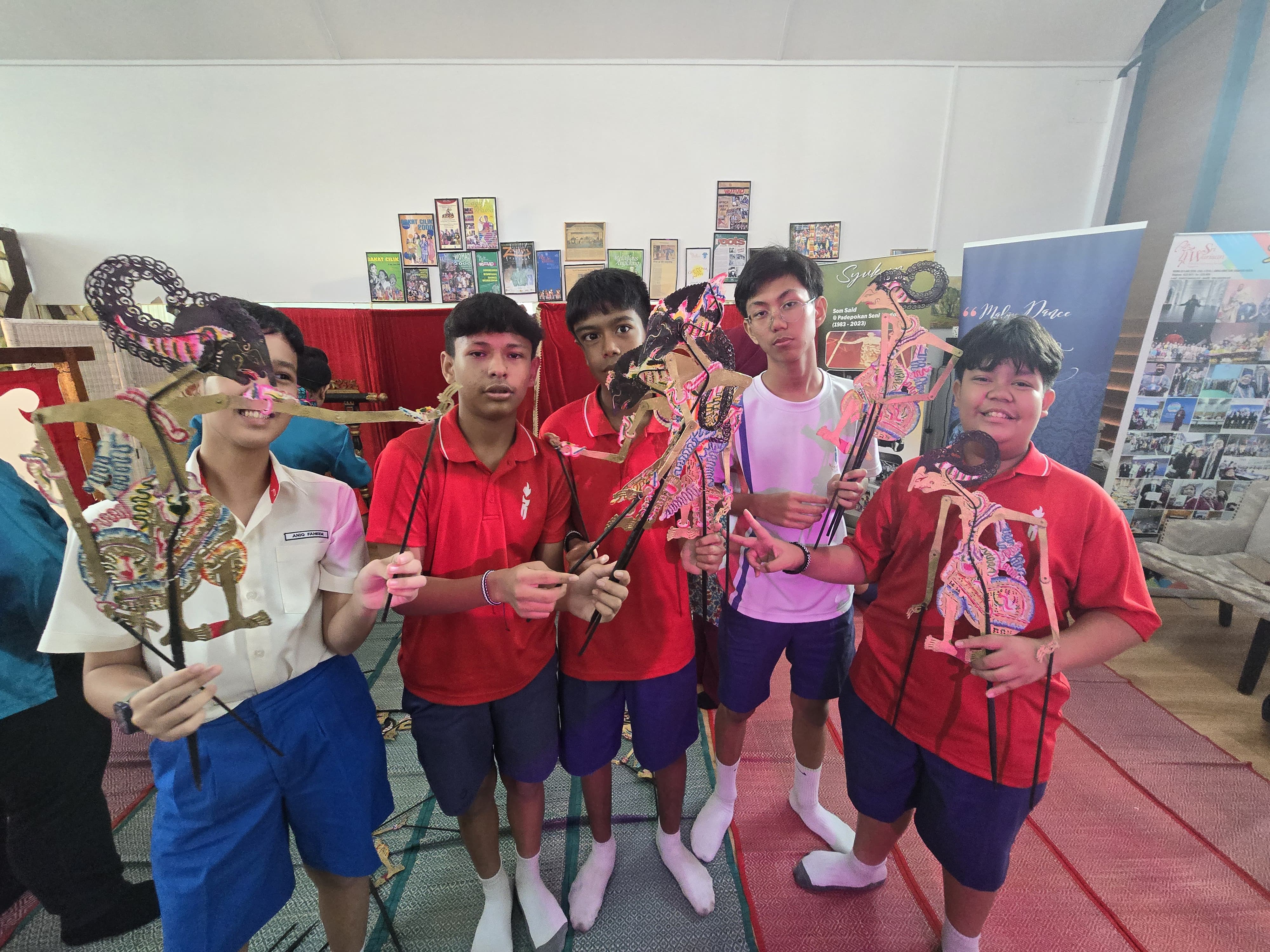 Five students in school uniforms hold colorful traditional wayang kulit shadow puppets indoors.