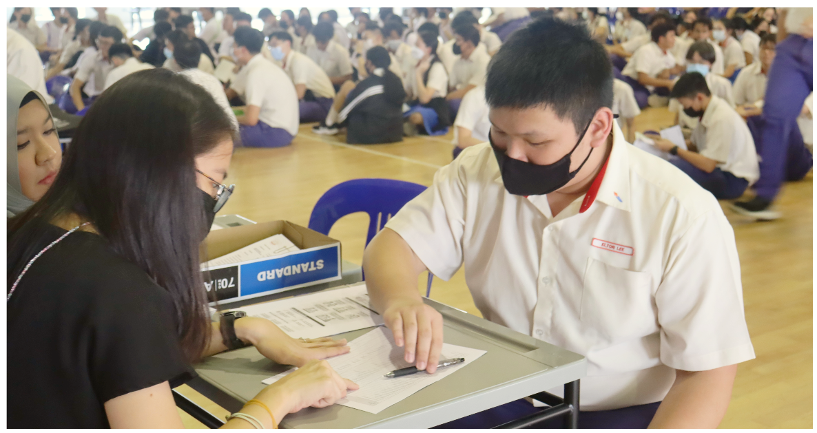 A masked student in uniform leans over a table reviewing papers with an adult in a crowded school hall.