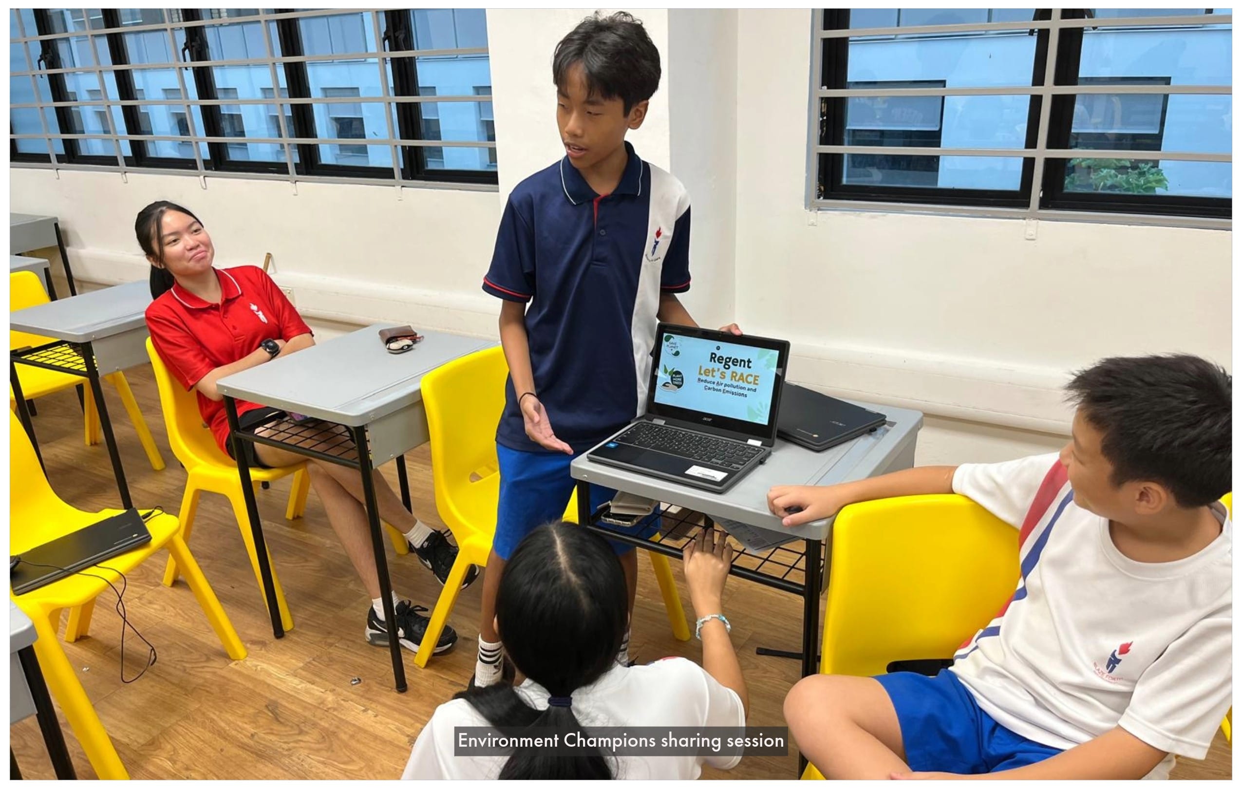 A student stands presenting from a laptop showing "Regent Let's RACE" to three seated peers in a classroom.