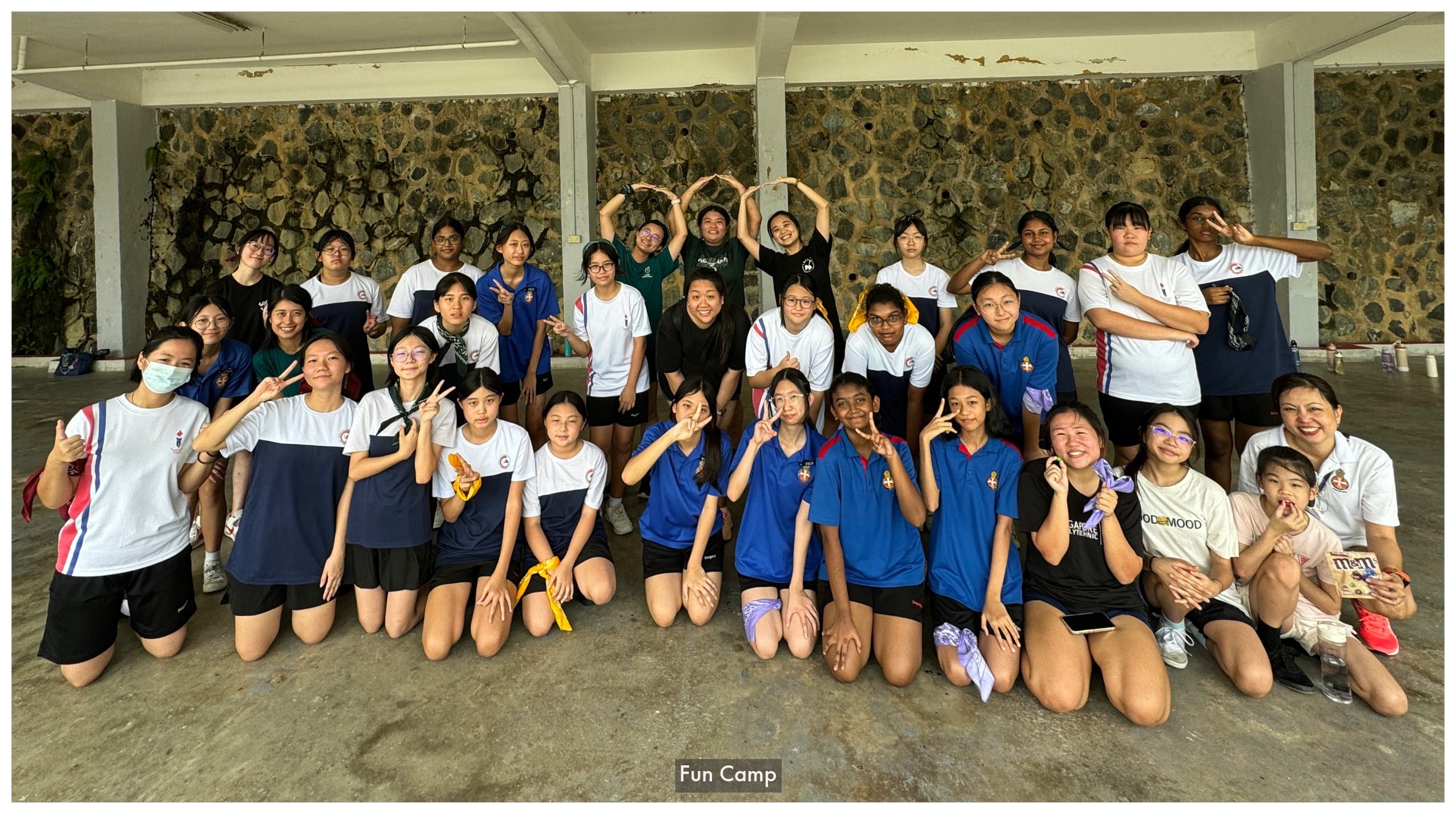 A large group of young women in sportswear pose together indoors at a fun camp, making peace signs and heart shapes.
