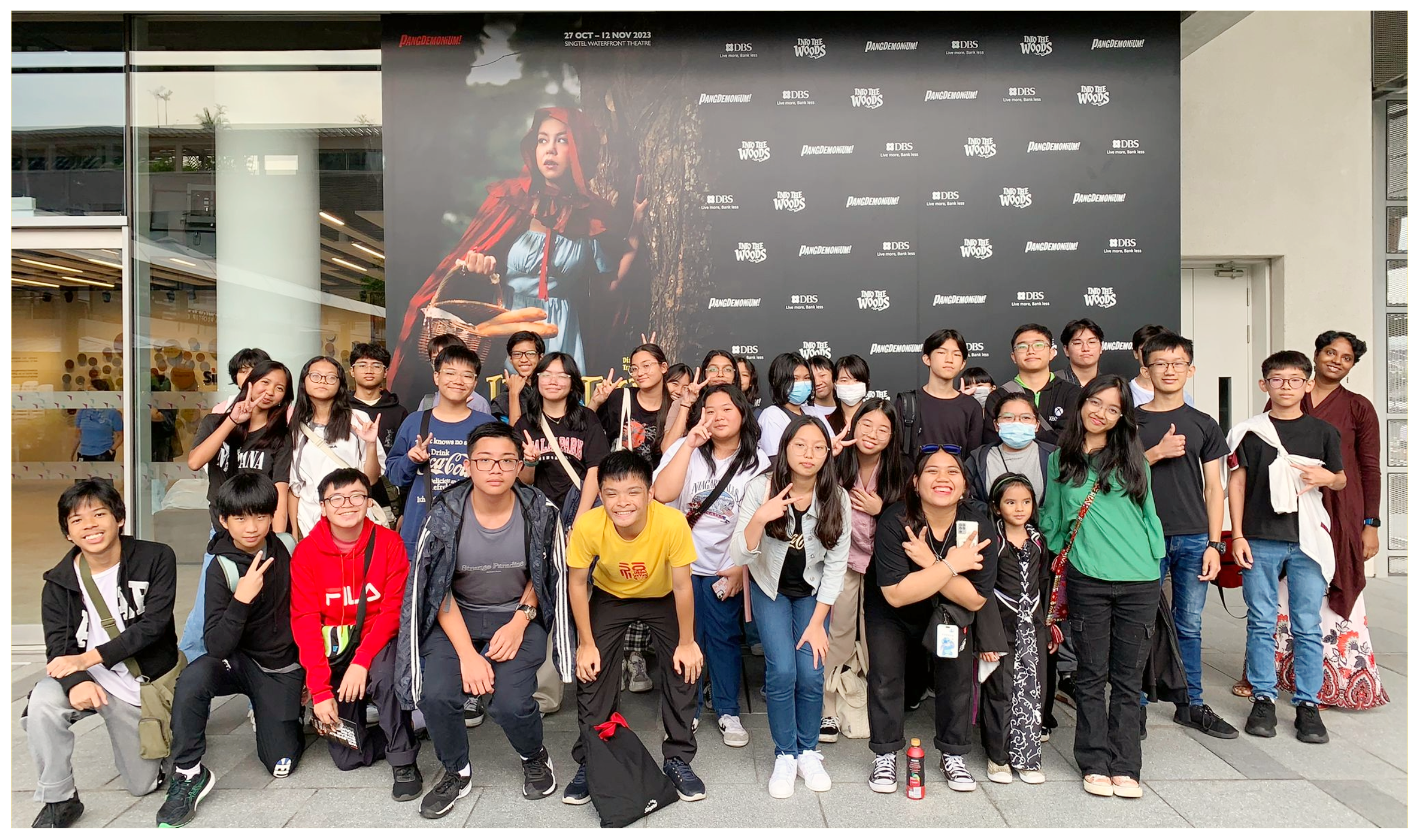 Large group of students posing in front of a theater backdrop featuring a girl in a red hood by a tree