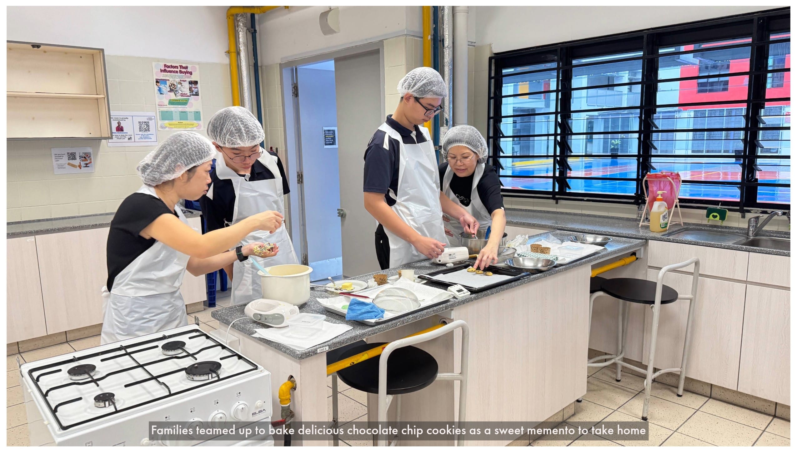 Four people wearing hairnets and aprons prepare cookies on a kitchen counter in a school cooking classroom.