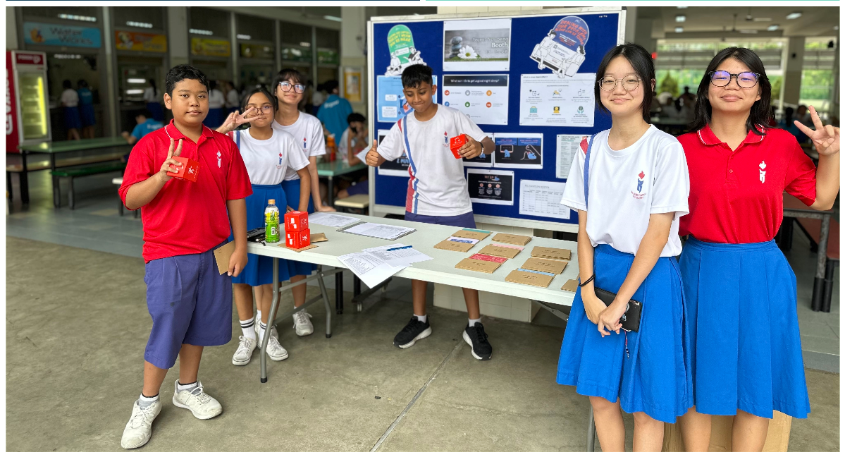 Six school students in uniforms stand at a project booth with a display board and table covered in papers and boxes.