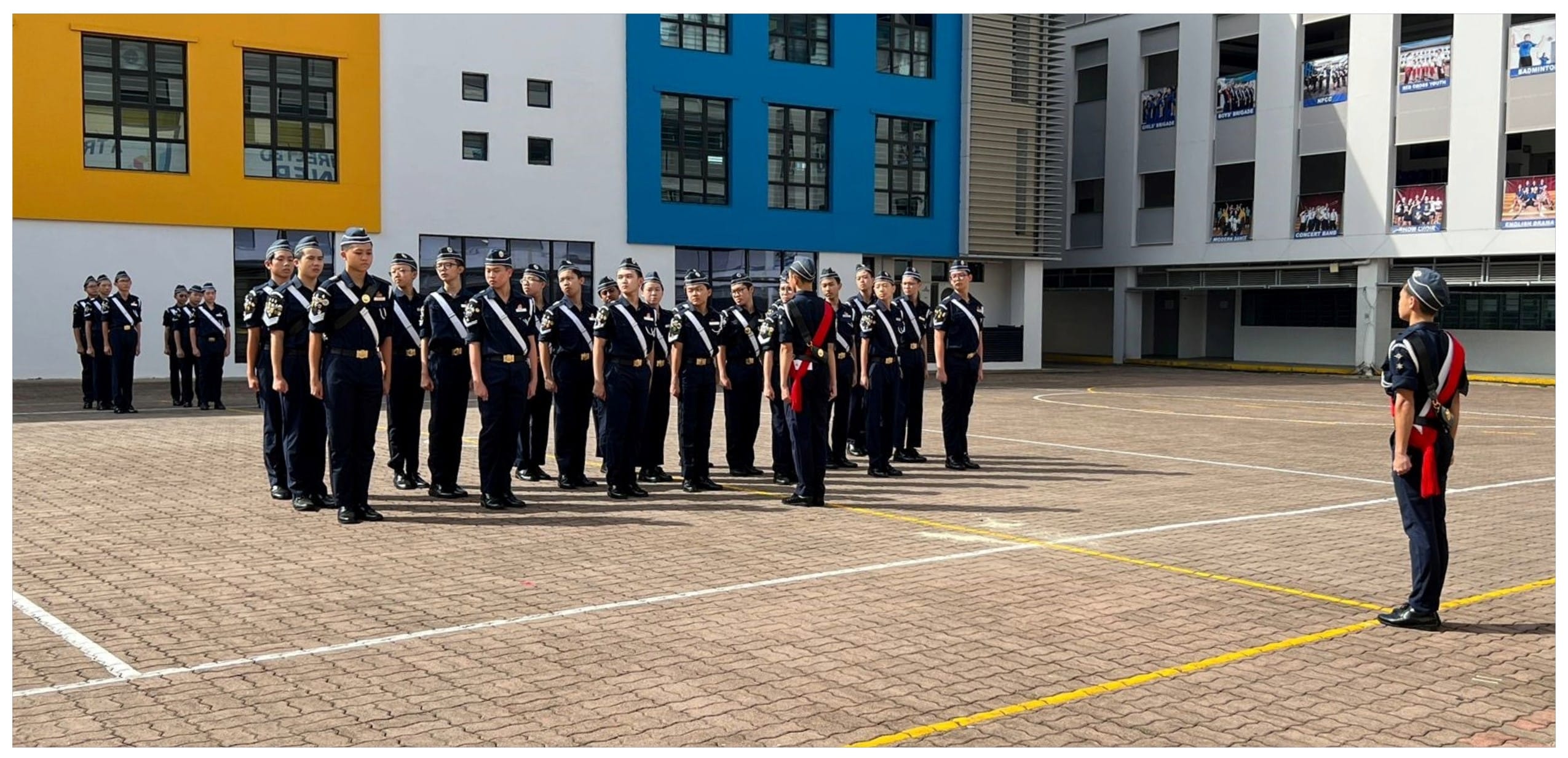 Cadets in dark navy uniforms stand in formation on a school courtyard while an officer faces them.