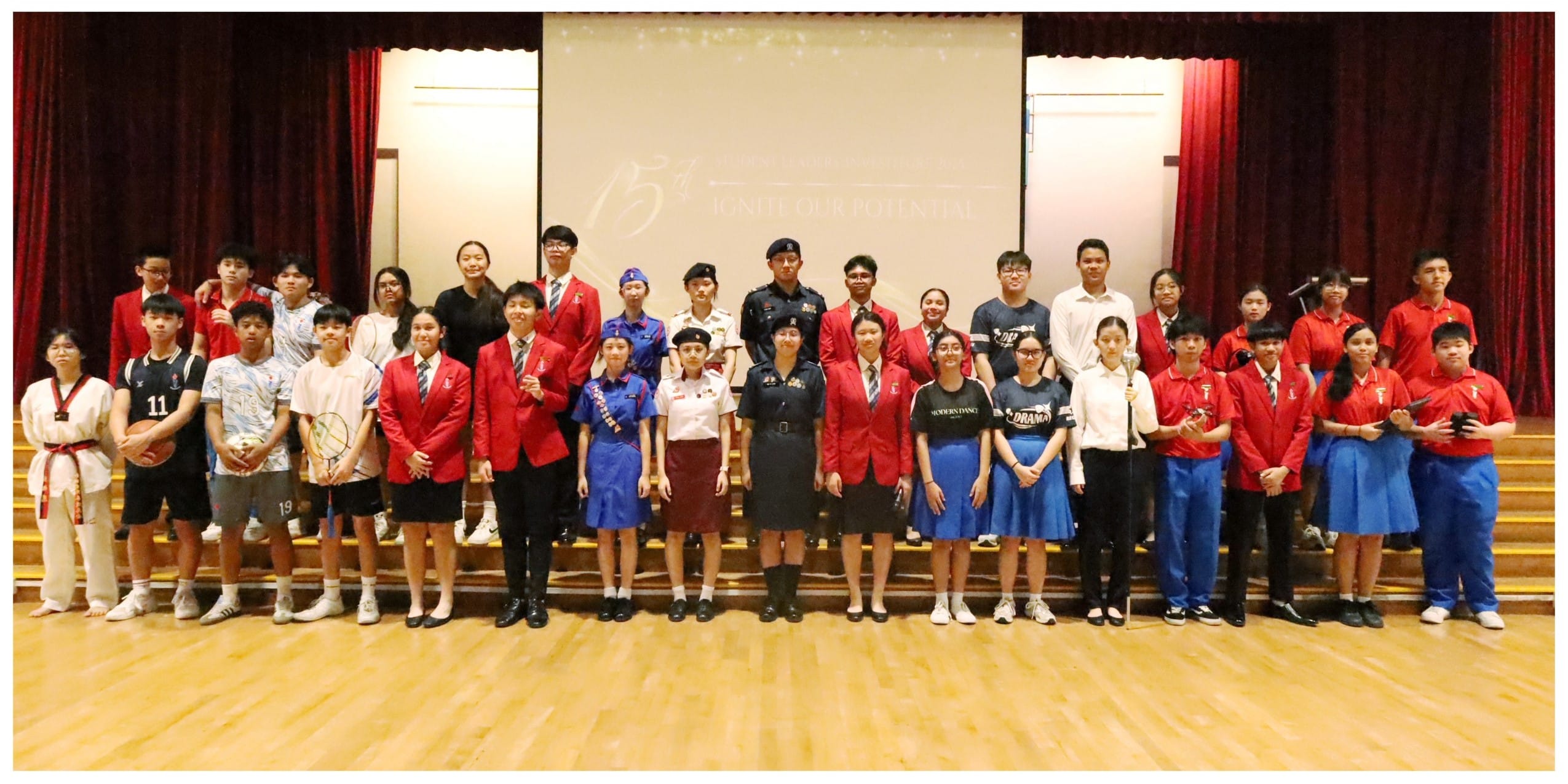 Large group of students in various uniforms and sports attire posing on a stage with a projection screen behind them.
