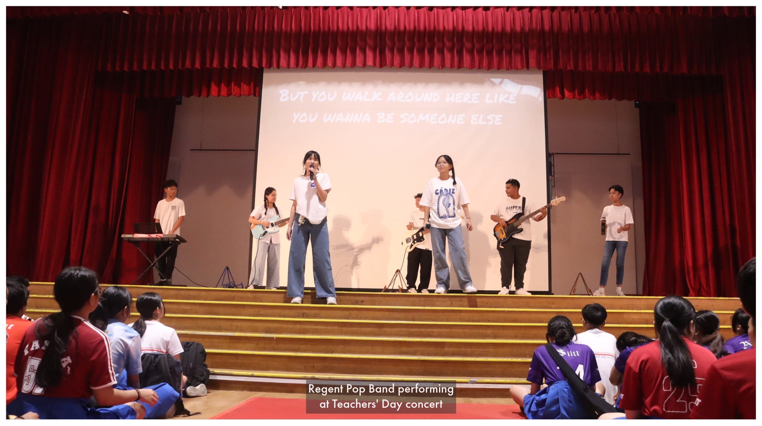 Students perform on a red-curtained stage with guitars and keyboard while an audience watches at a Teachers' Day concert.