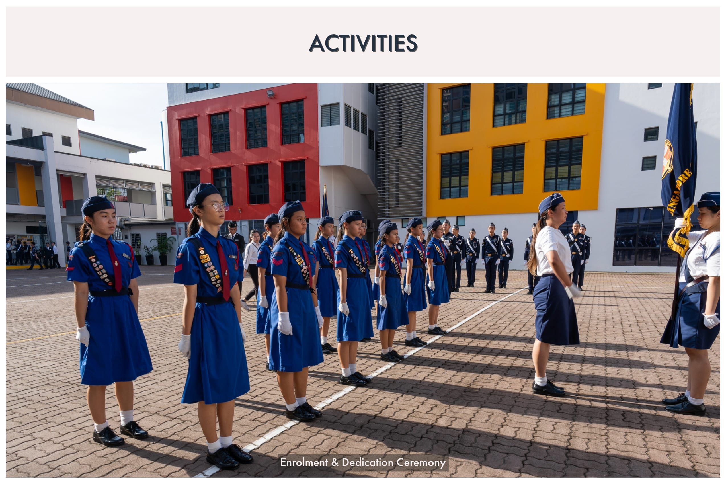 Students in blue uniforms with badges stand at attention during an Enrolment and Dedication Ceremony on a school courtyard.