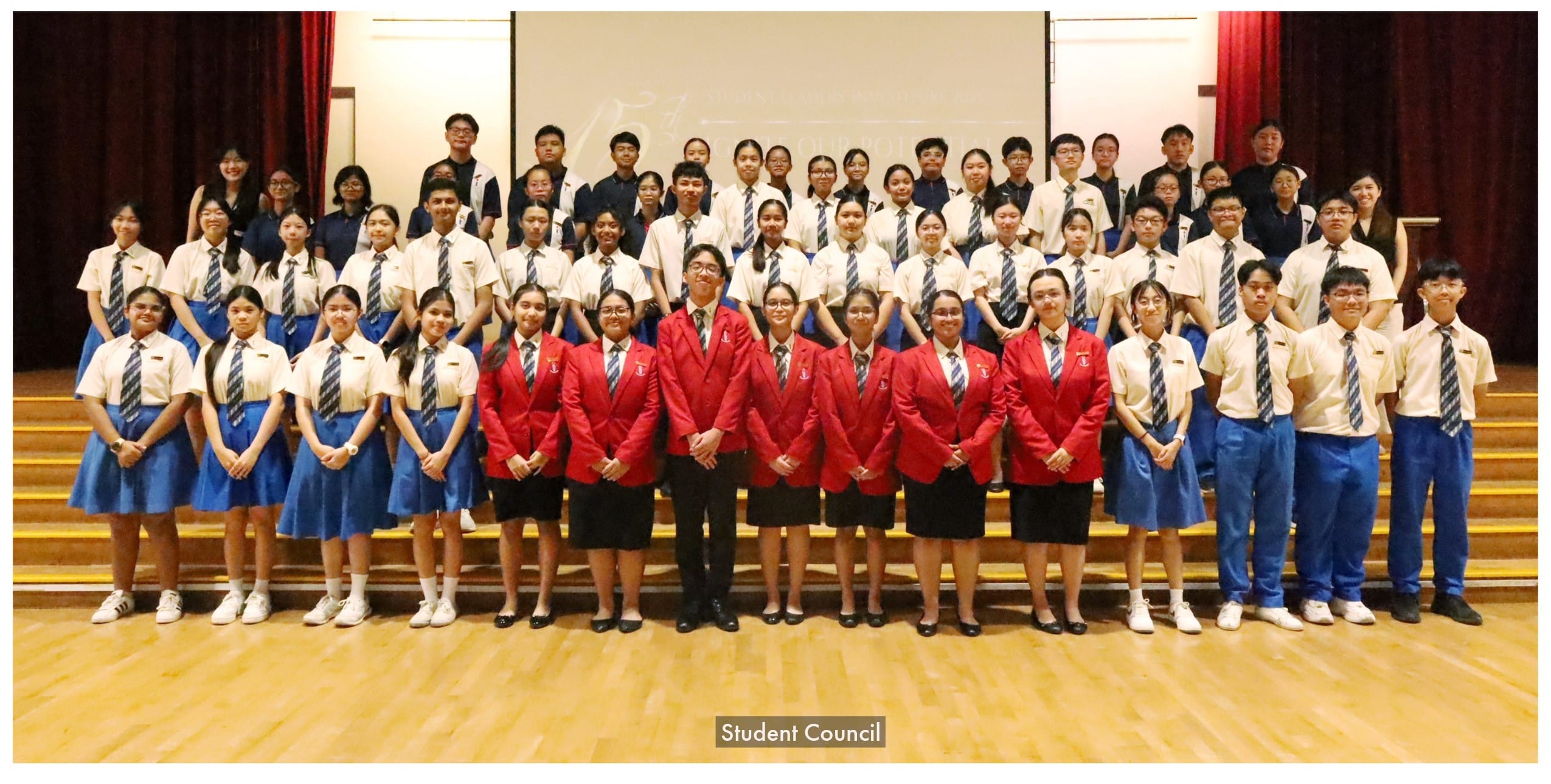Large group of uniformed students posing on a stage, with leaders in red blazers standing in the front row.