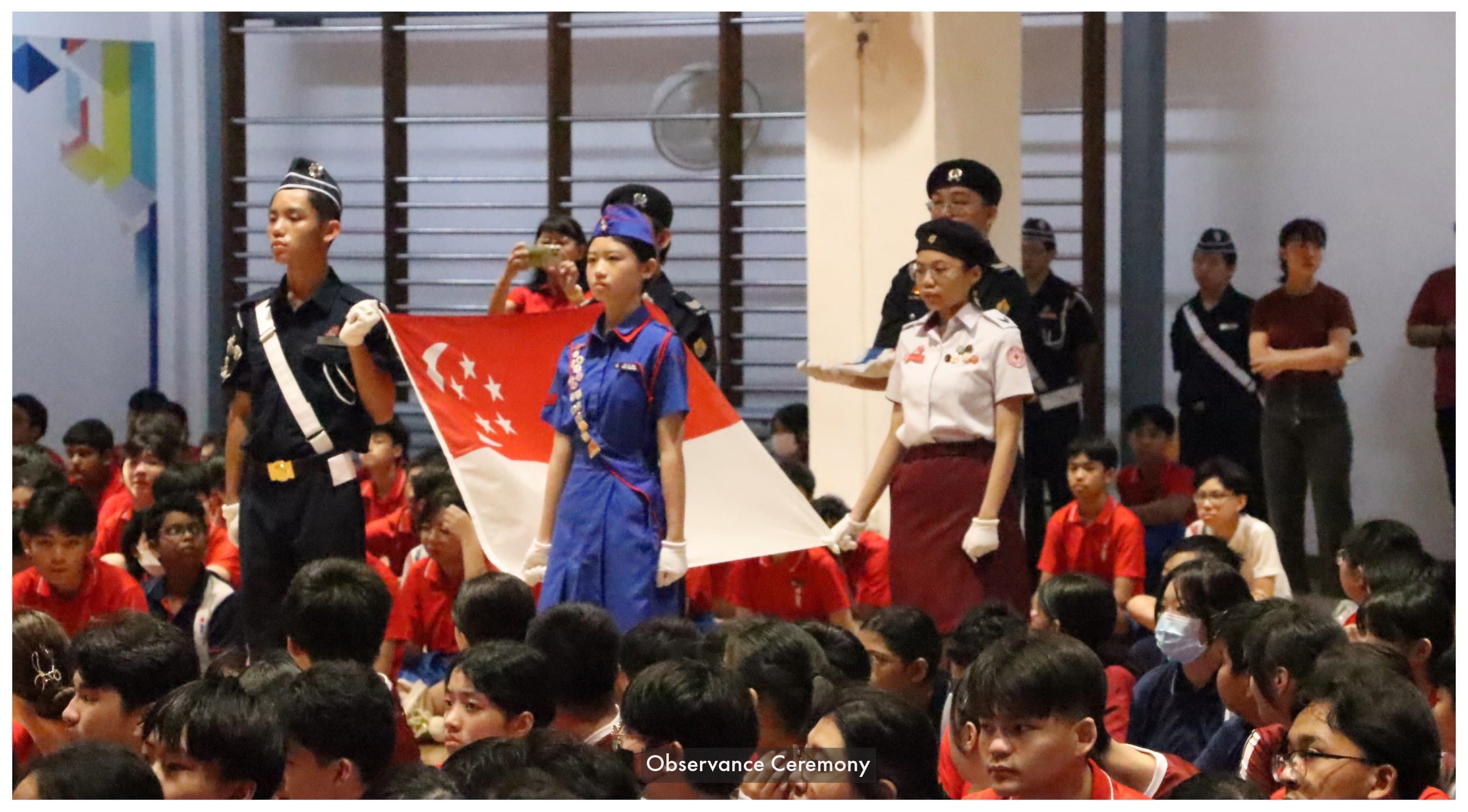 Students in uniform carry the Singapore flag during an observance ceremony in a school hall filled with seated pupils.
