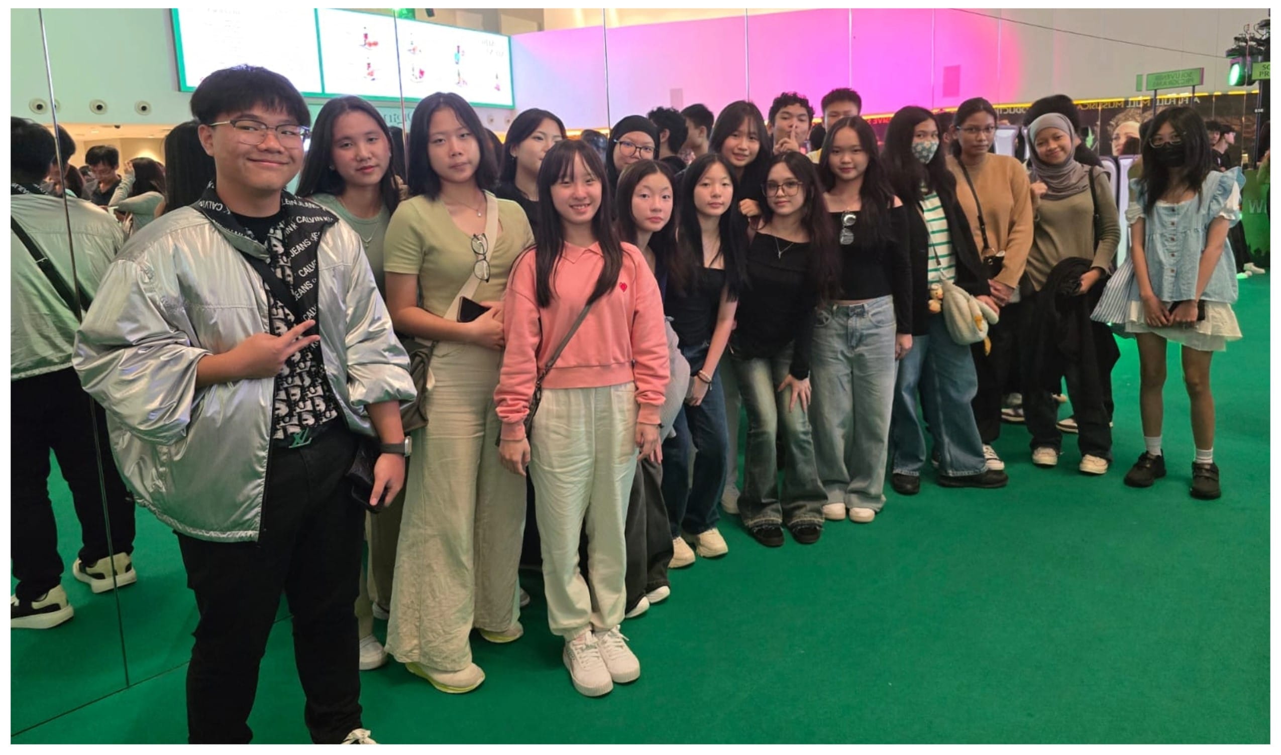 A large group of young people posing together on a green floor inside a brightly lit venue with pink lighting.