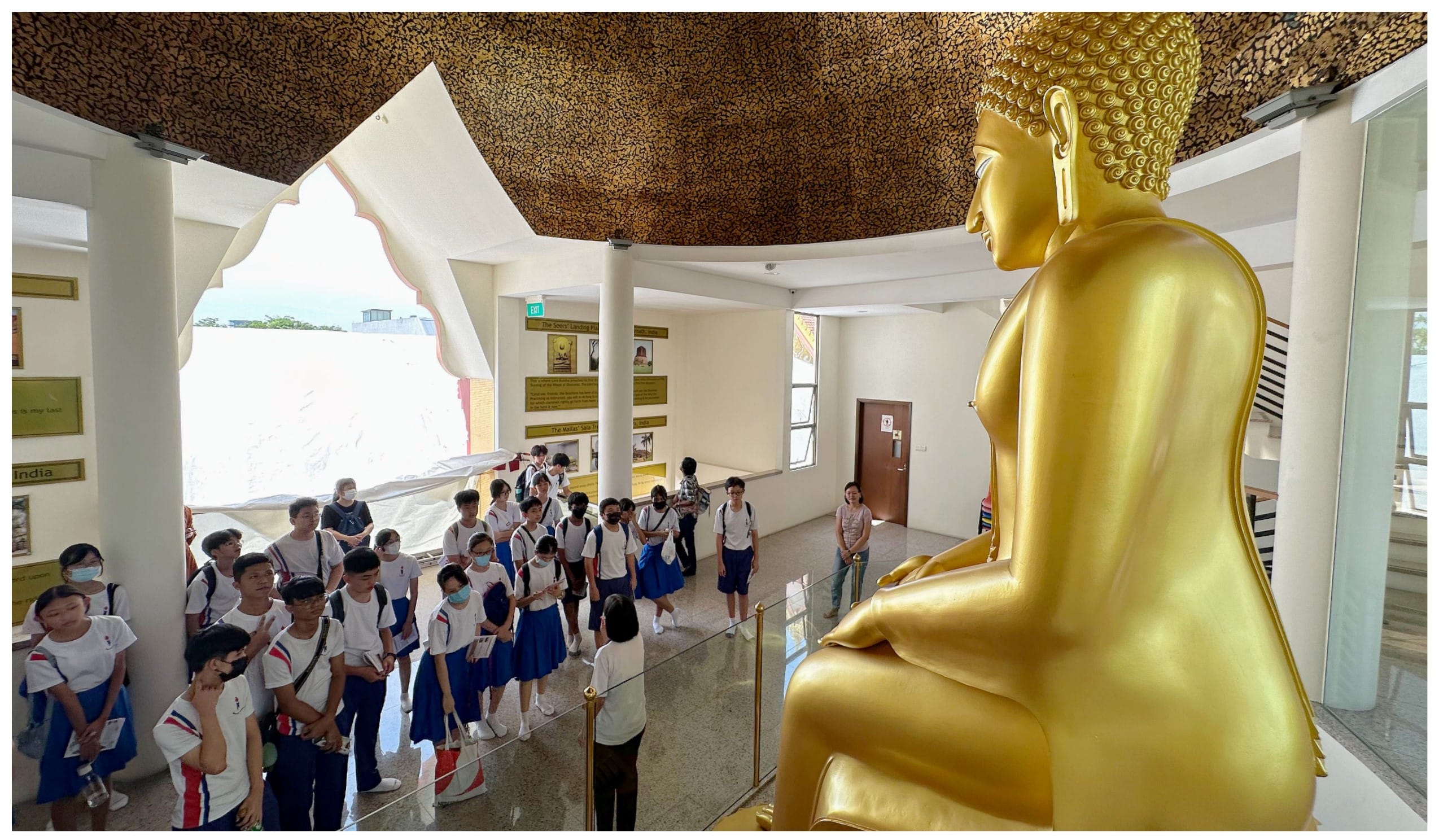 A group of school children in uniforms stand inside a temple hall before a large golden seated Buddha statue.