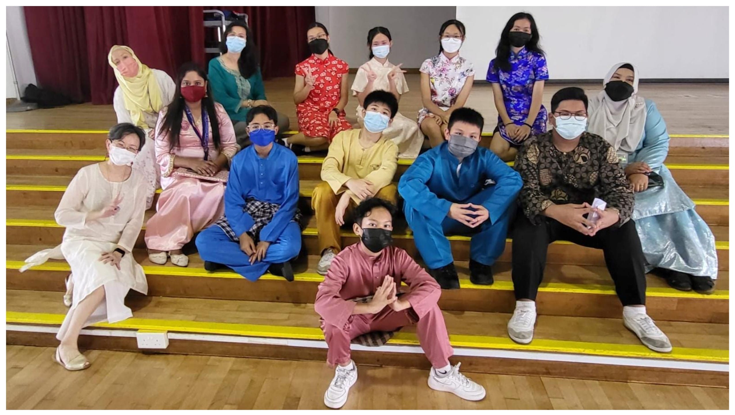 A group of masked students in colorful traditional outfits sit on wooden bleachers in a hall.