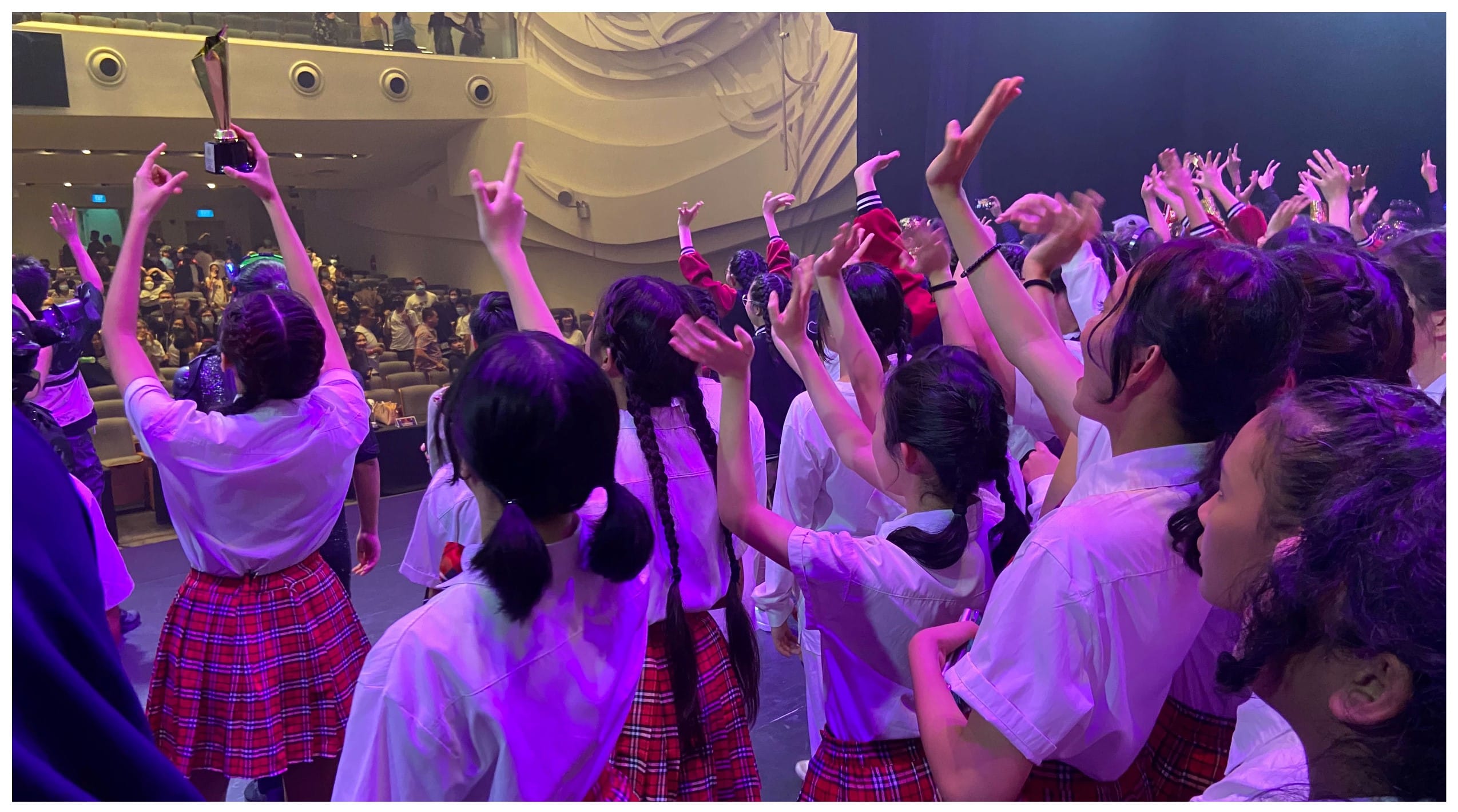 Girls in white shirts and red plaid skirts raise hands on a purple-lit stage, one holding a trophy.