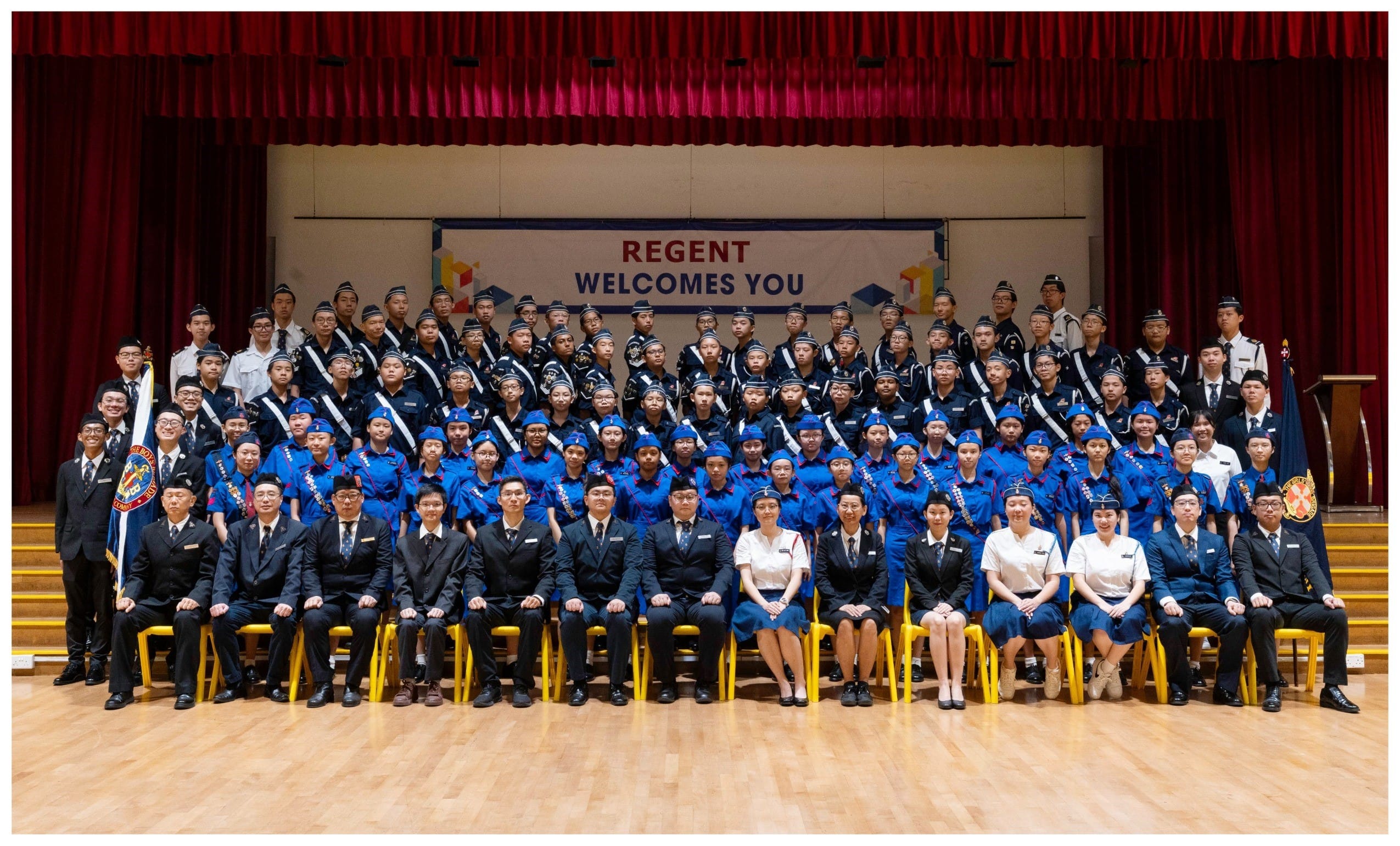 Large group of uniformed students and staff posed on a stage in front of a Regent Welcomes You banner.