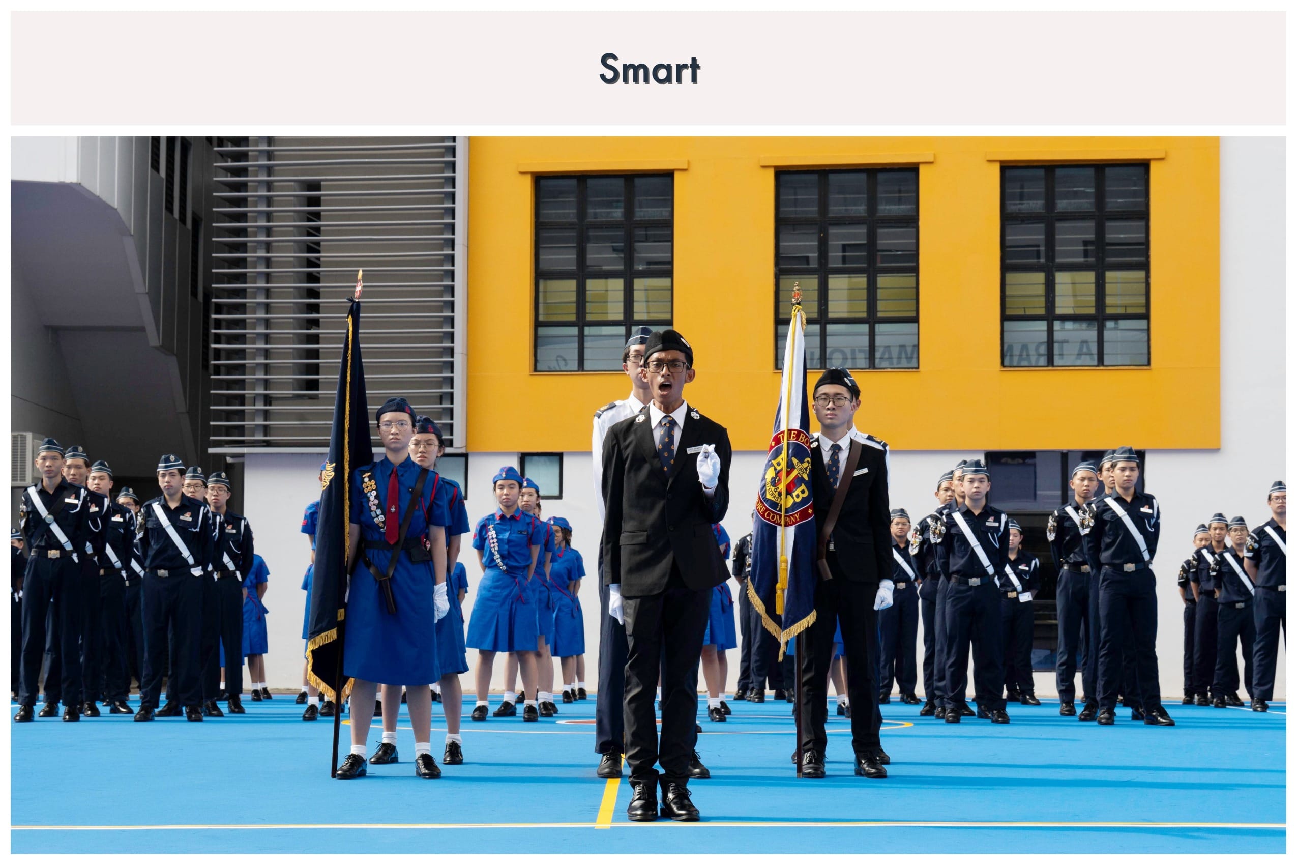 Students in uniform stand at attention during a formal parade on a blue court in front of a yellow building.