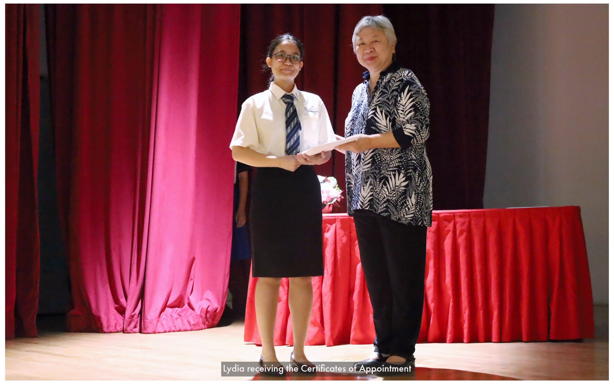 A young student in uniform receives a certificate from an older woman on a stage with red curtains.