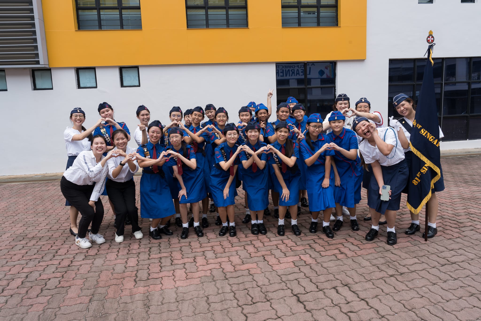 A large group of girls in blue uniforms and caps pose making heart shapes with their hands outside a school building.
