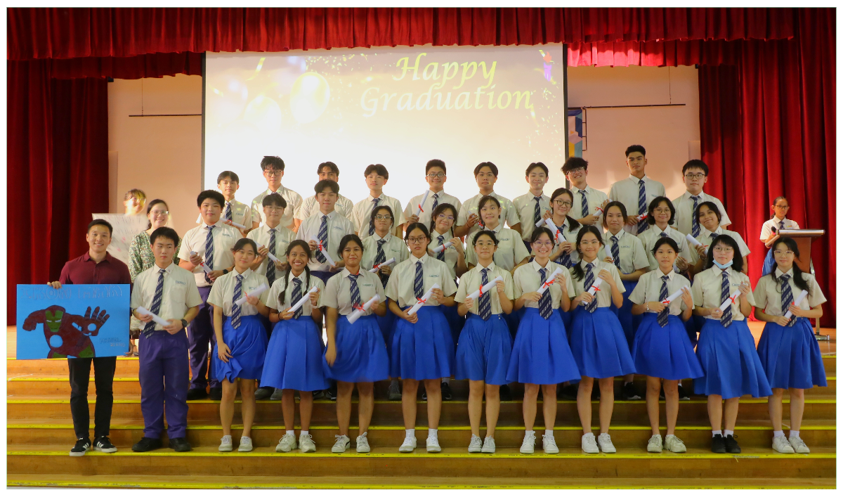 A large group of students in uniforms holding diplomas on a stage with a Happy Graduation screen behind them.