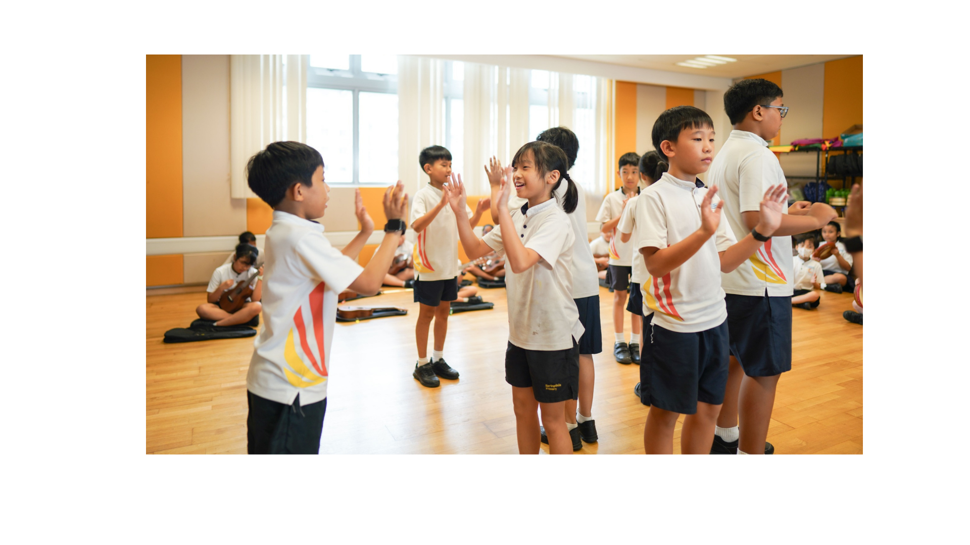 Primary school children in white uniforms clapping hands with partners in a bright indoor activity room.