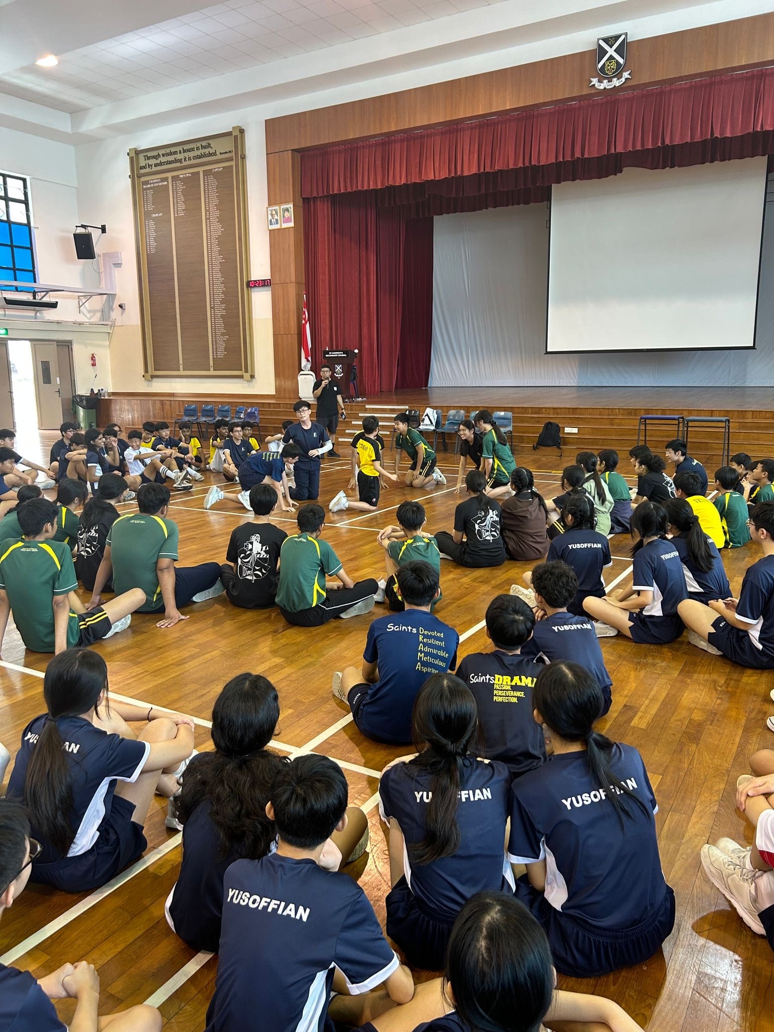 Students in navy and green uniforms sit on a school hall floor facing a speaker near a stage with red curtains.