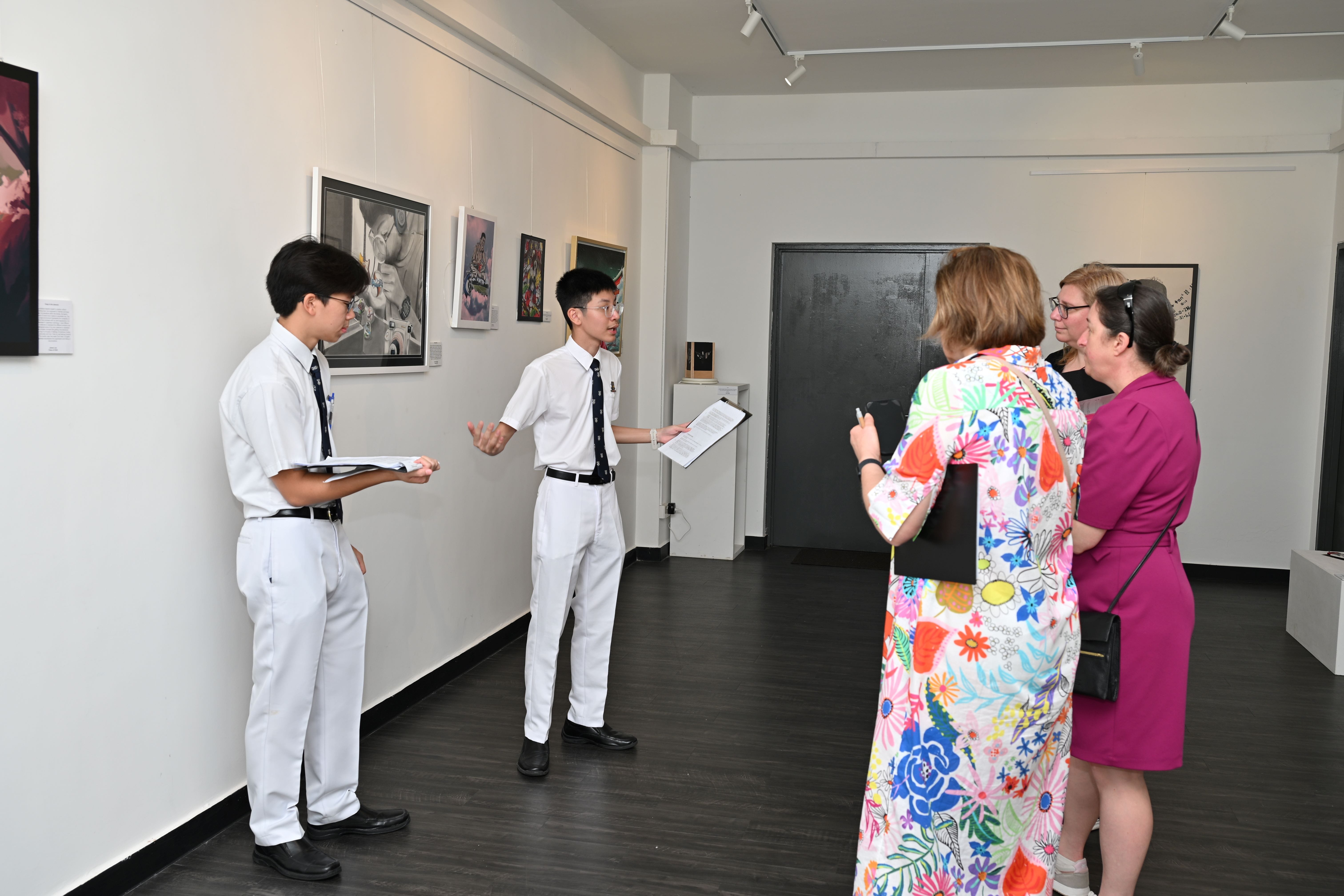 Two students present artwork to three adult women in a white gallery room with framed pieces on the walls.