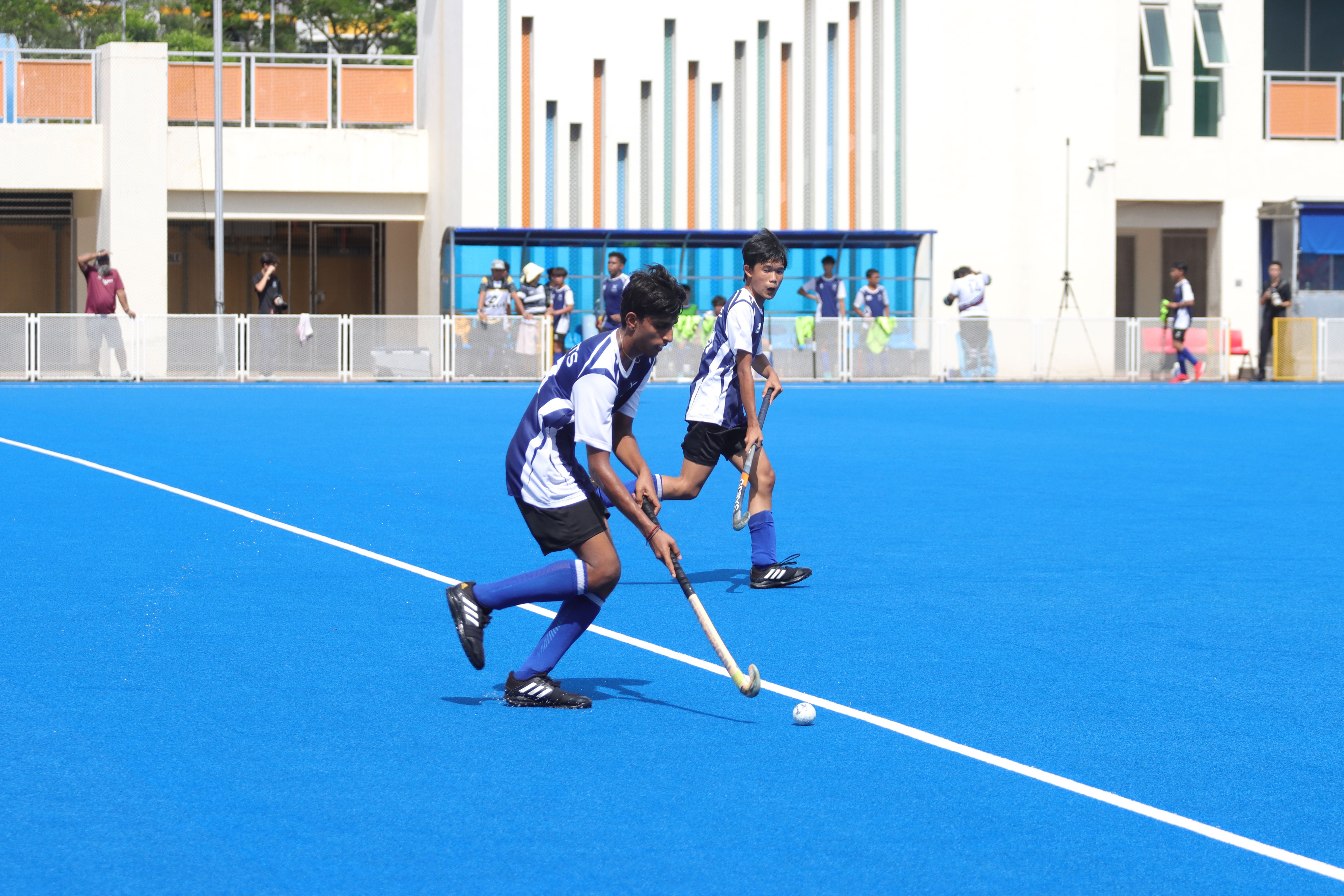 Two young boys in blue and white uniforms play field hockey on a bright blue turf court.