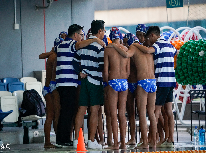 A water polo team in blue swim caps and trunks huddles with coaches in striped shirts beside an indoor pool.
