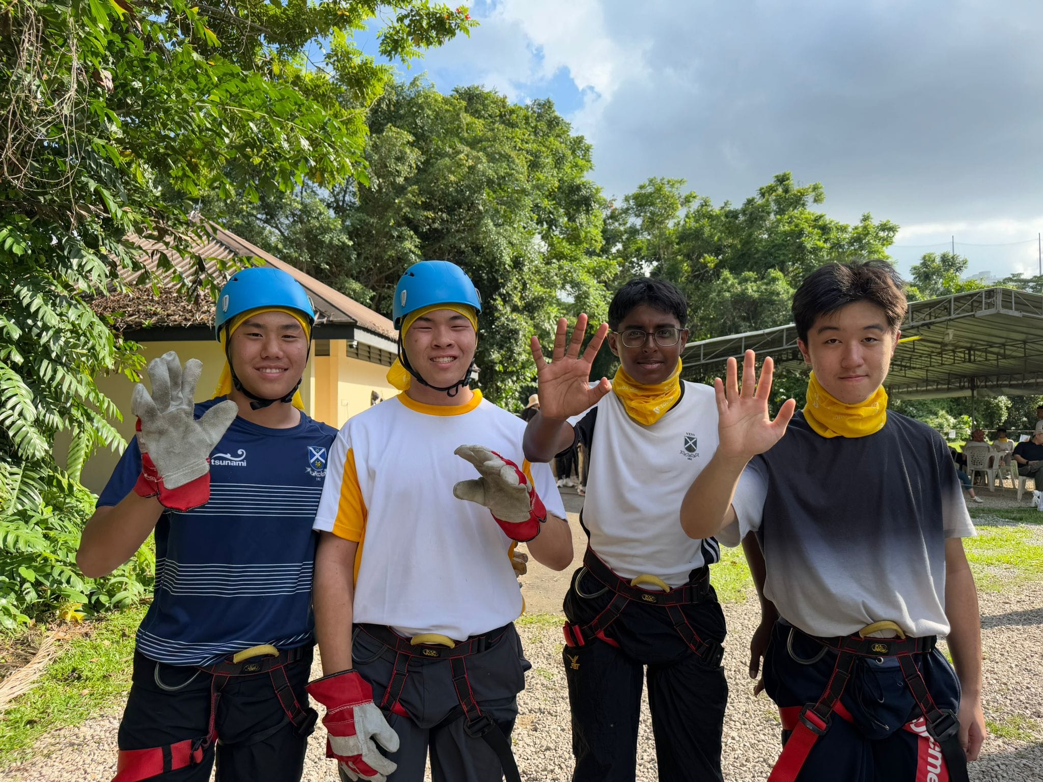 Four boys in safety harnesses and helmets wave at the camera outdoors near trees.