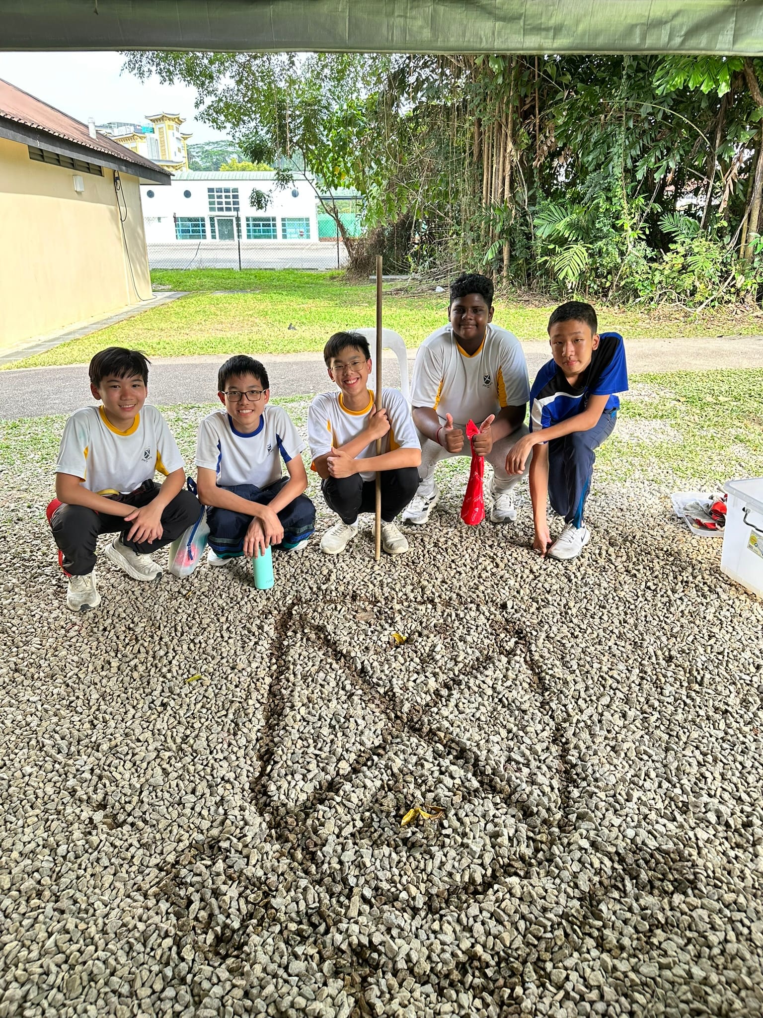 Five boys in school uniforms crouch on a gravel surface outdoors, smiling