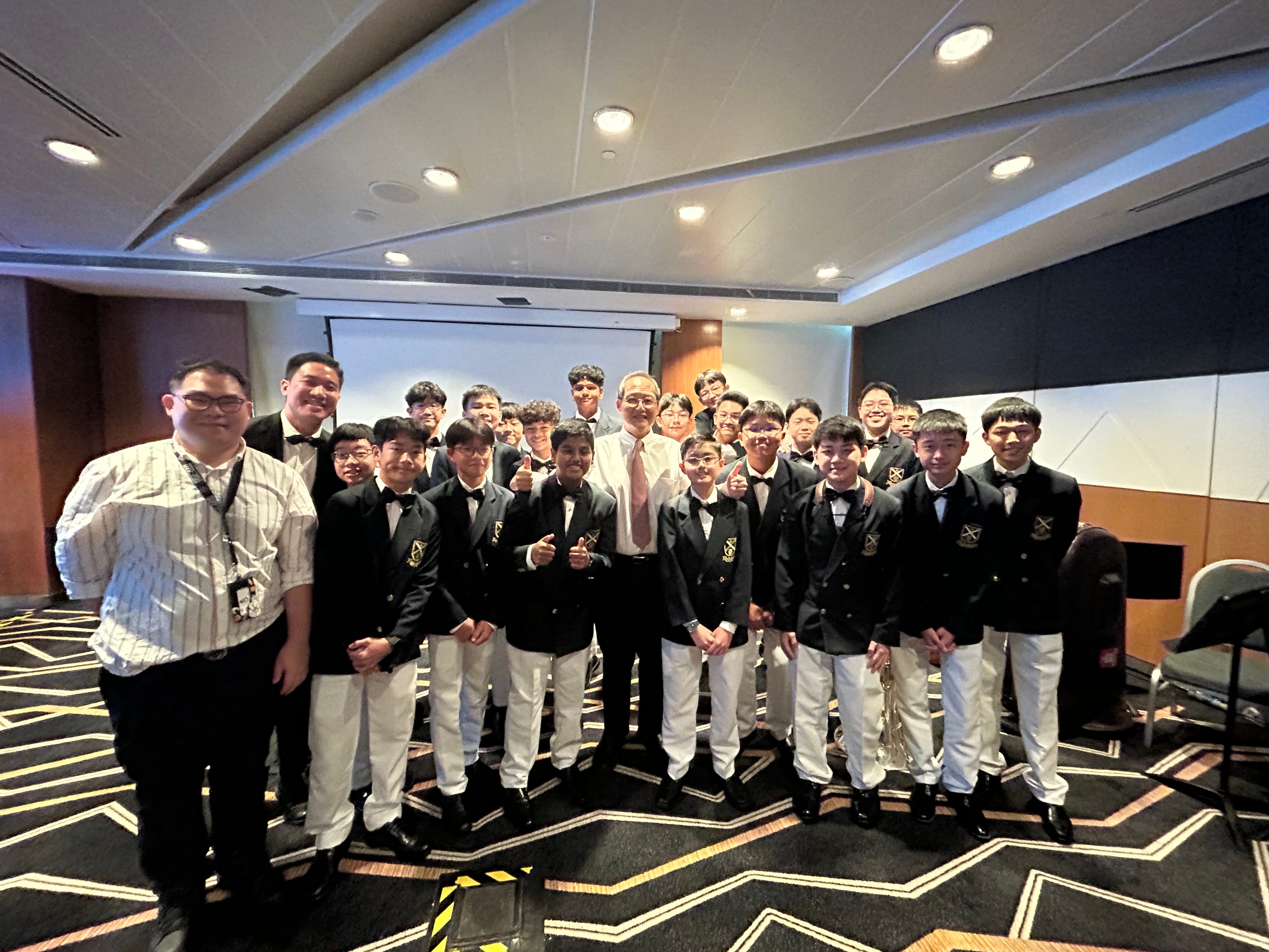 A large group of young males in black blazers and white trousers pose with adults in a formal indoor room.