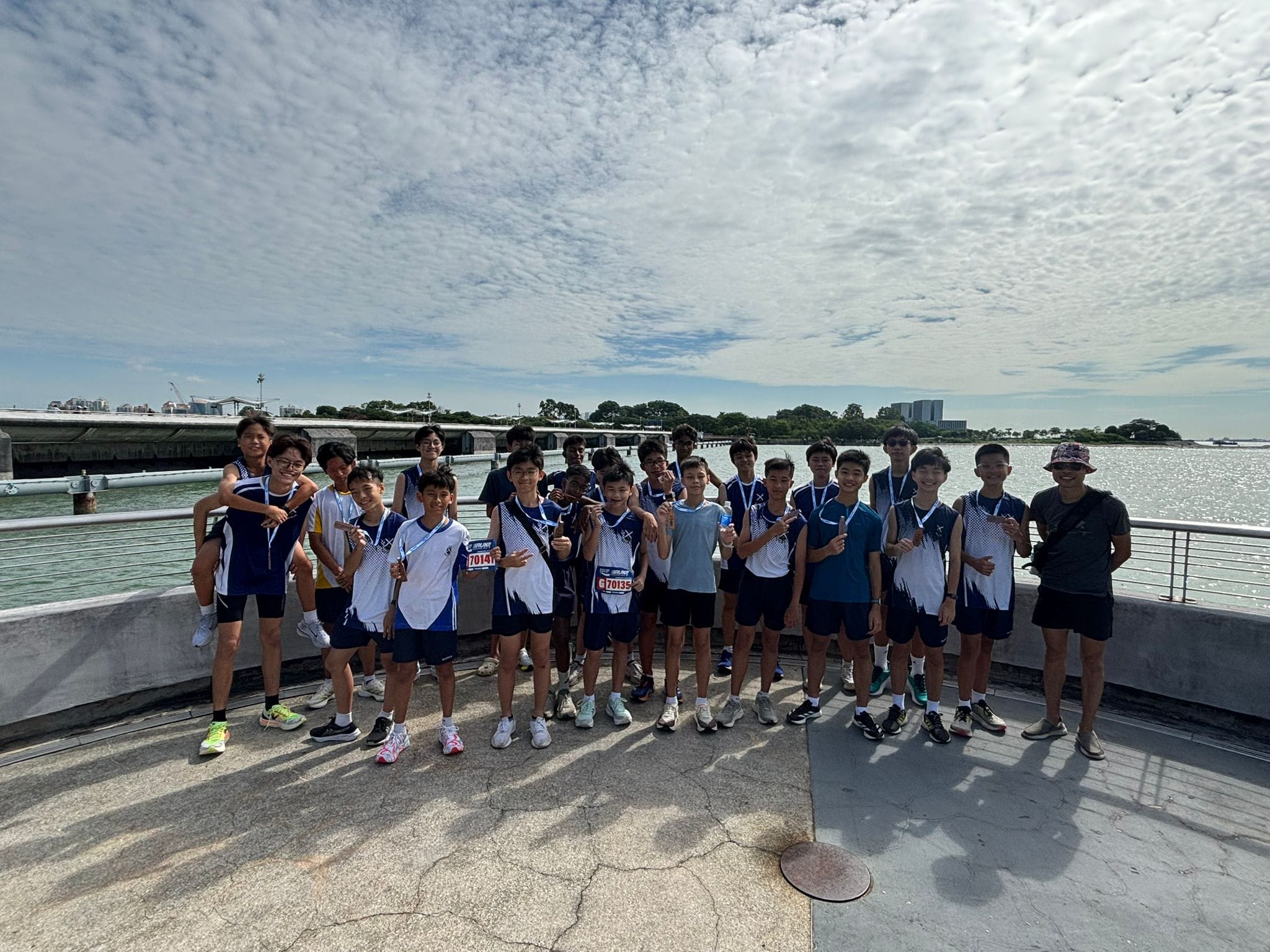 A group of young runners in blue and white uniforms pose with medals on a waterfront under a cloudy sky.