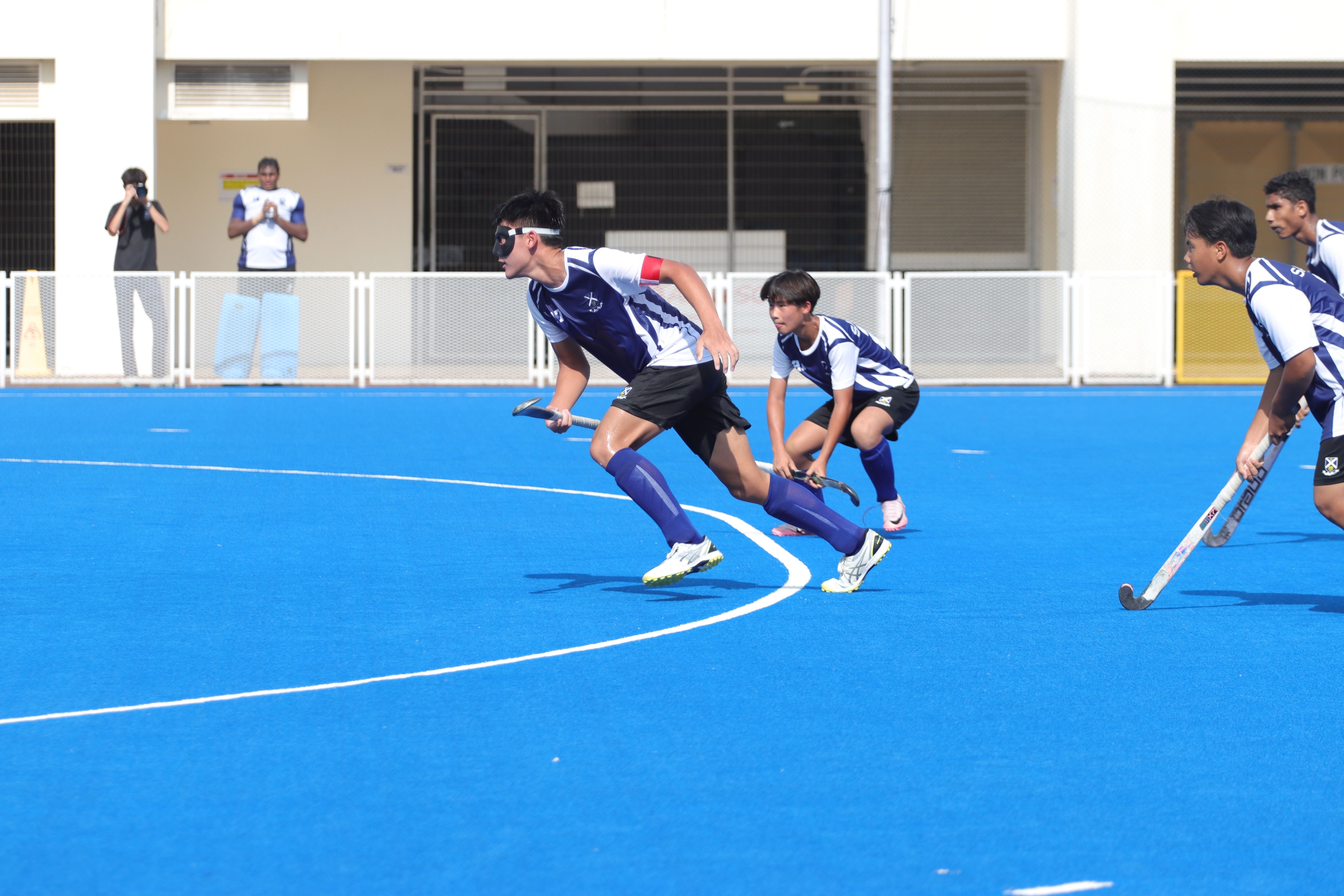Three teenage boys in blue and white uniforms play field hockey on a bright blue artificial turf court.