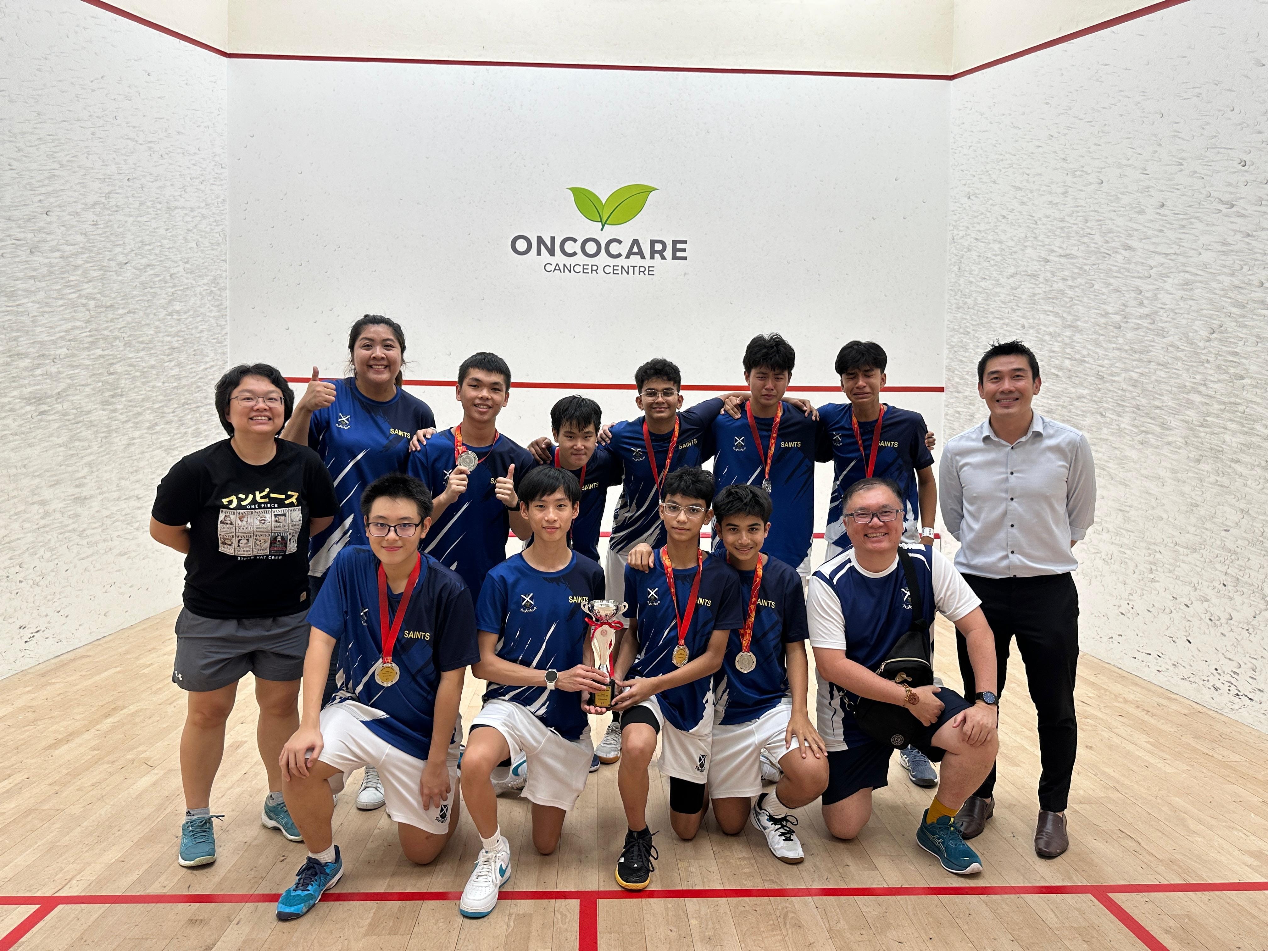 A youth squash team in blue Saints jerseys poses with medals and a trophy on an indoor court at Oncocare Cancer Centre.