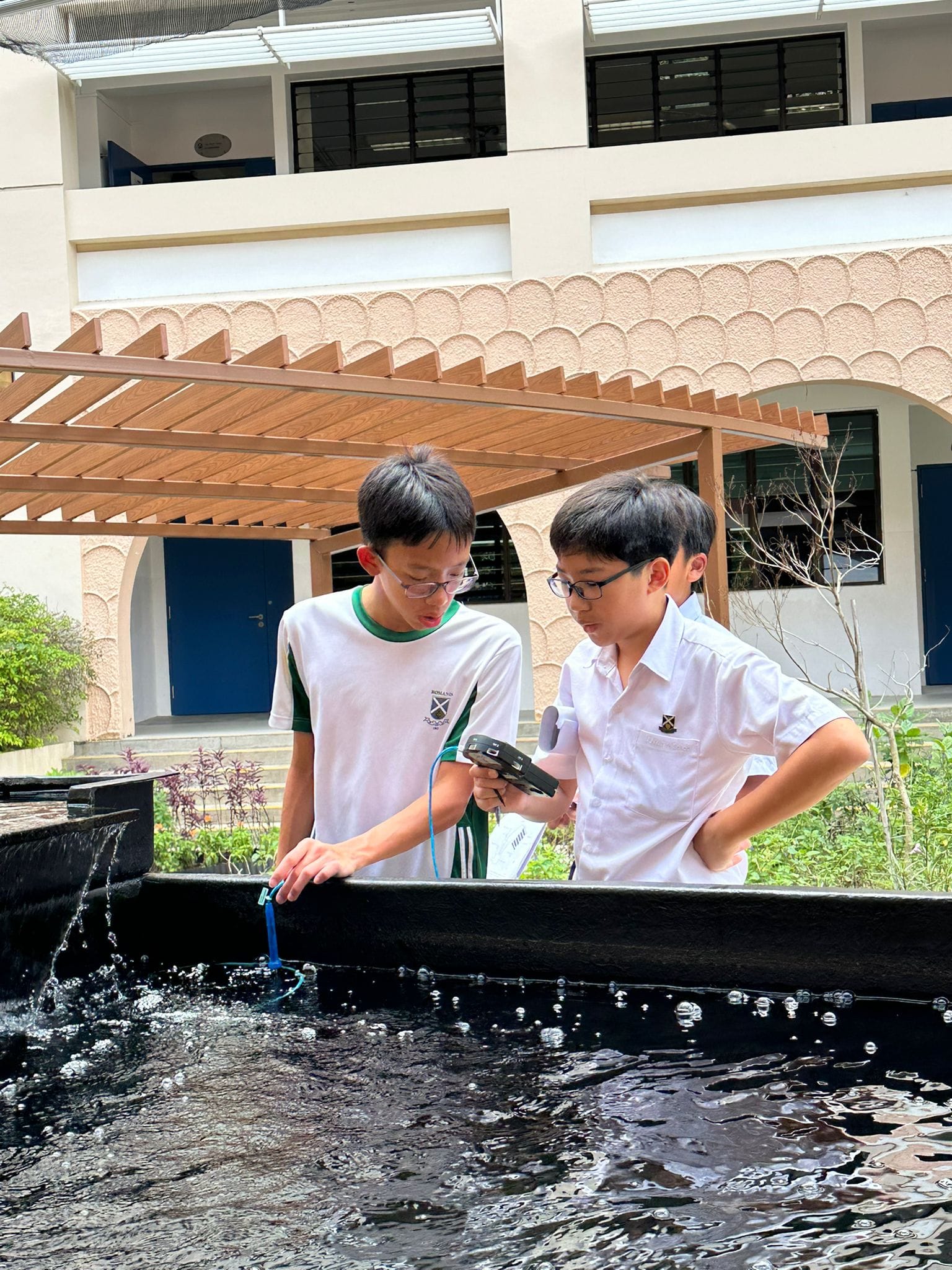 Two boys in school uniforms test water in an outdoor pond using a handheld device and a probe.