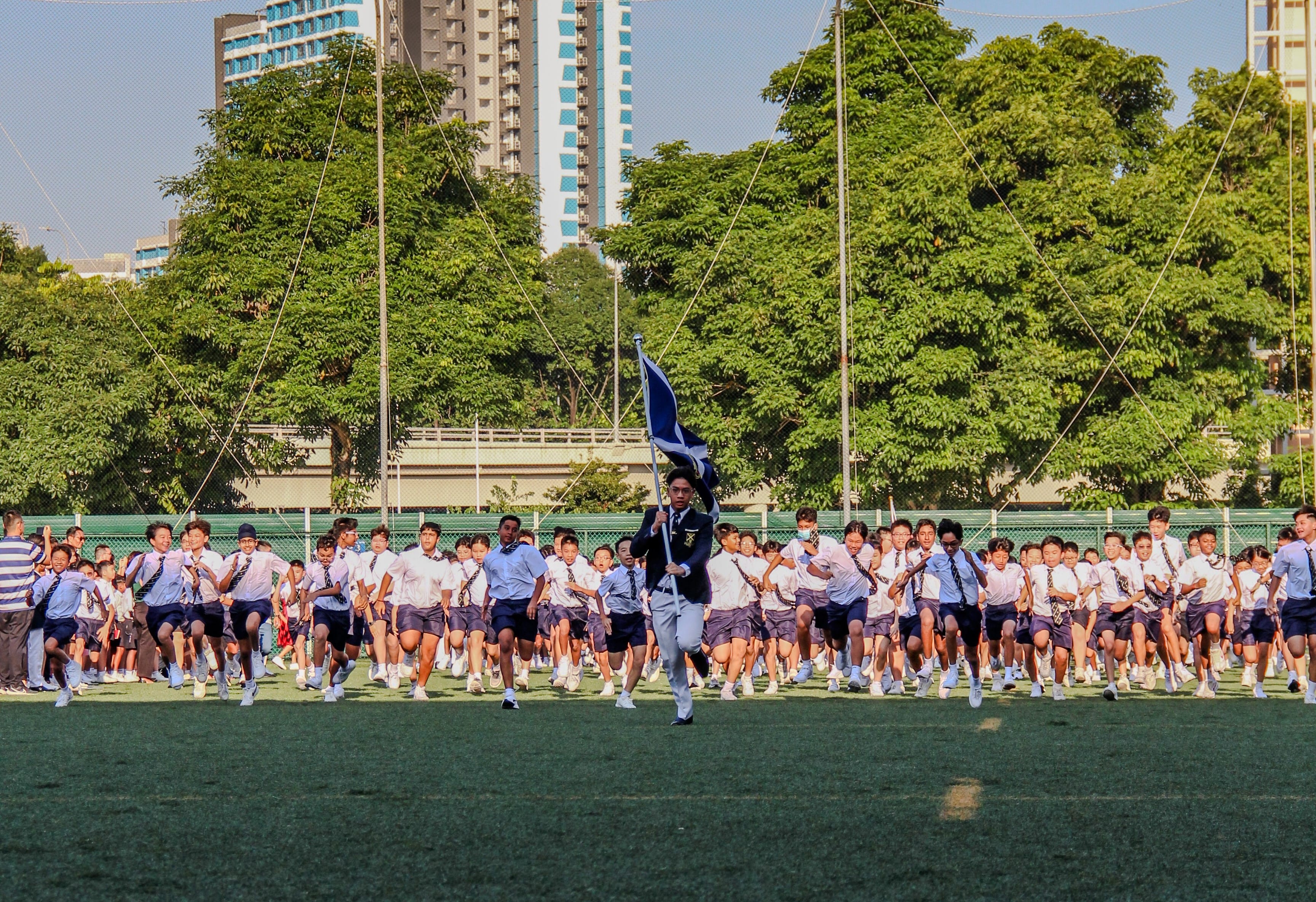 A large group of uniformed students runs across a green field, led by a student carrying a blue flag.