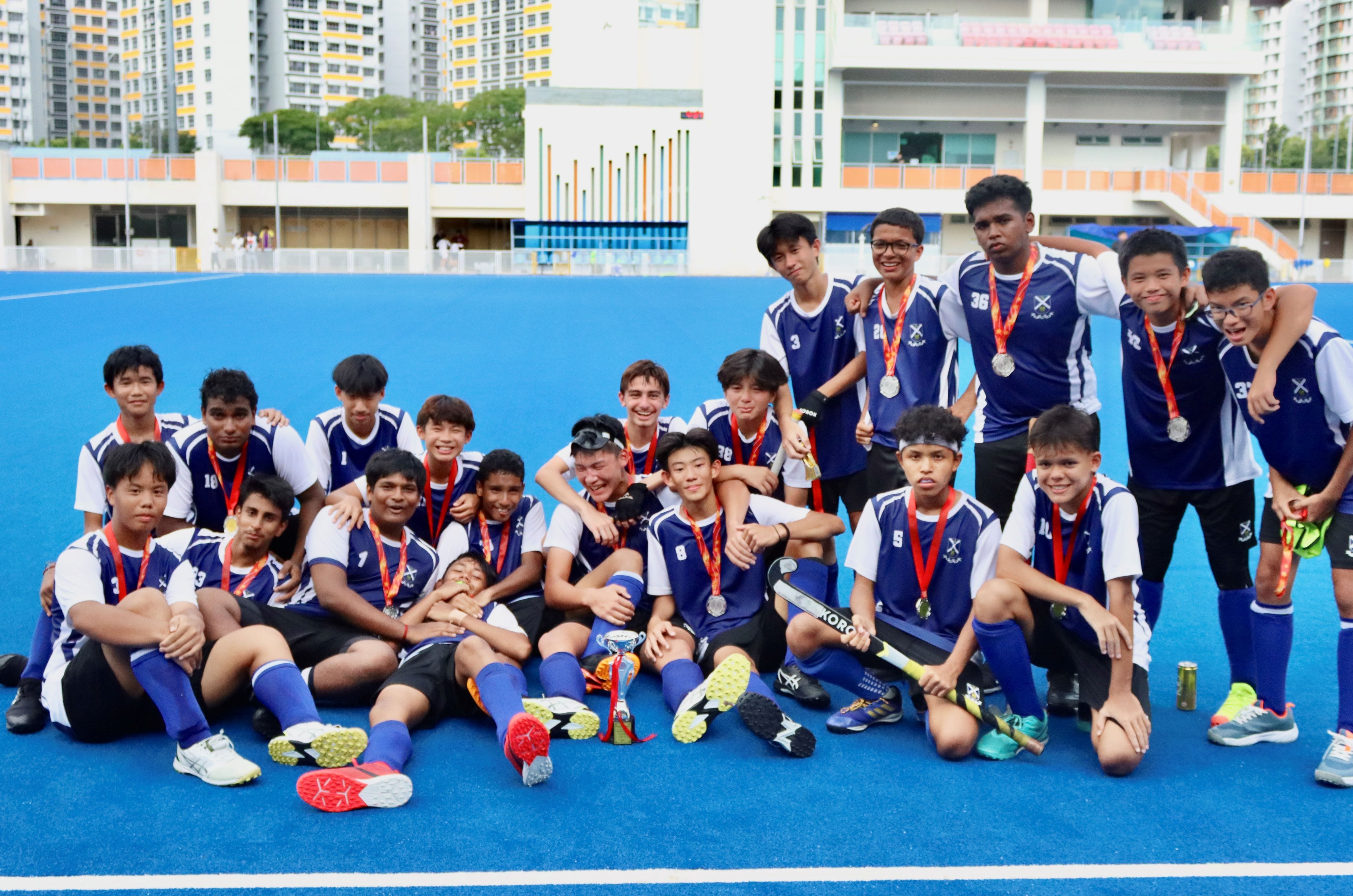 A youth field hockey team in blue uniforms poses with medals and a trophy on a blue turf field.