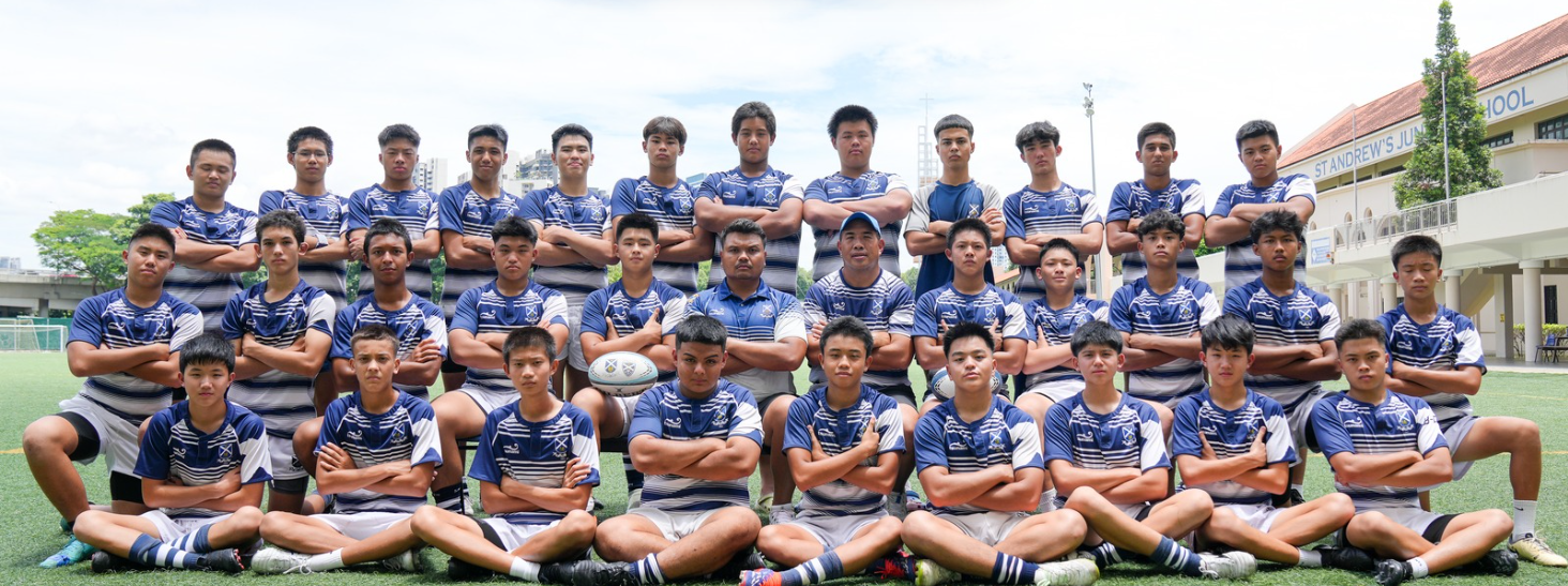 A youth rugby team in blue and white striped jerseys poses with arms crossed on a field near St Andrew's Junior School.