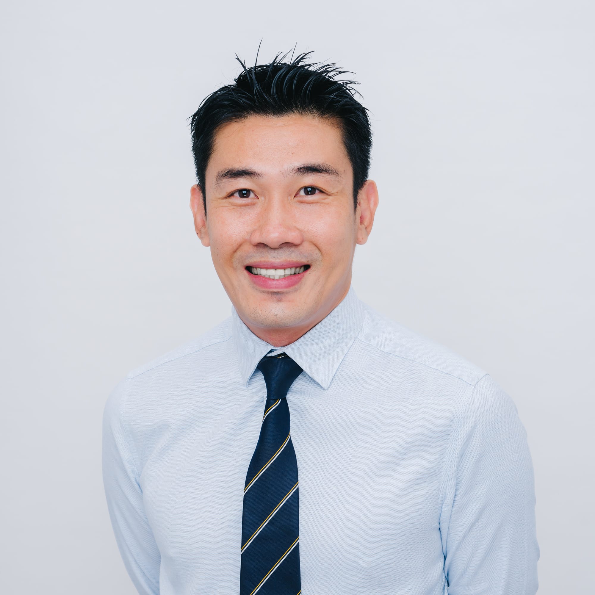A smiling man with short black hair wearing a light blue shirt and navy striped tie against a white background.